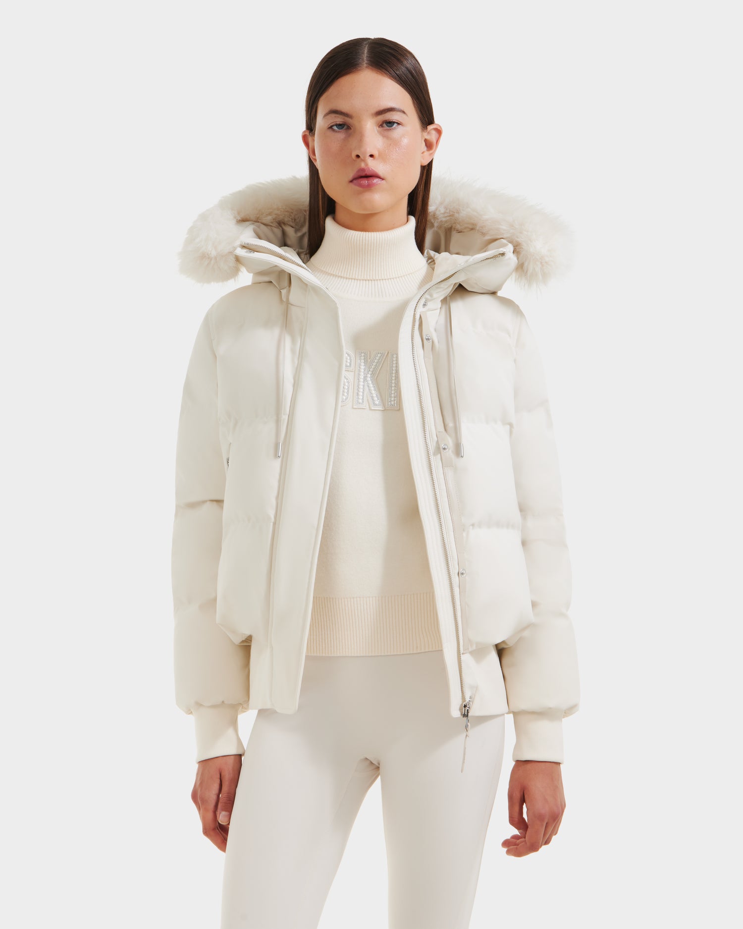 A woman faces forward against a plain white background, wearing the Rudsak CIEYNA - 8125579 Vanilla down-filled bomber with a faux fur hood, a cream turtleneck sweater, and white pants.