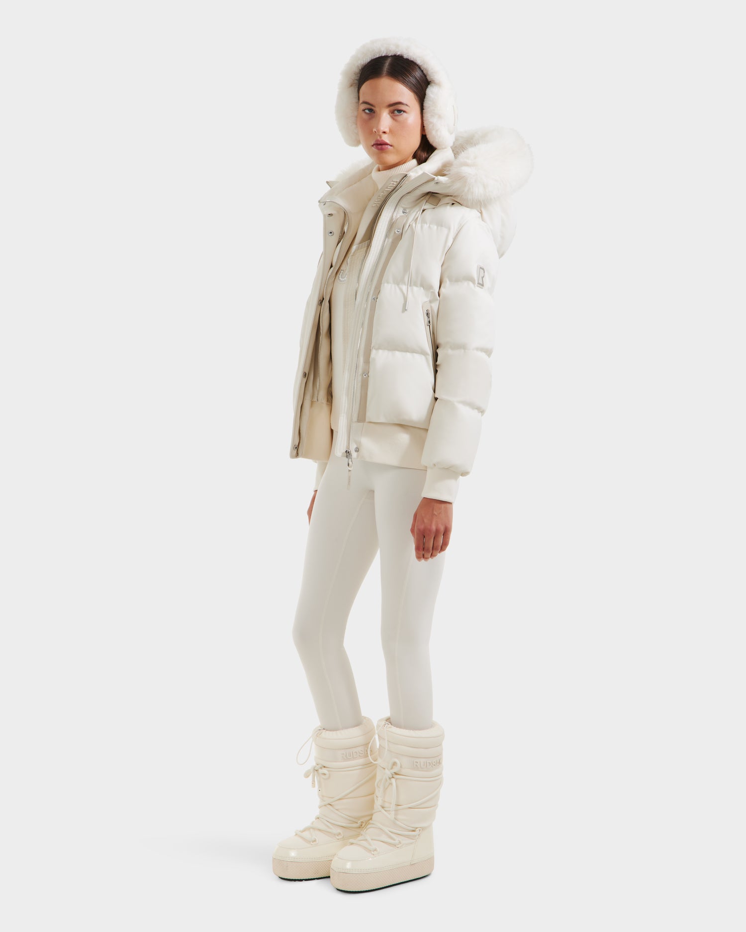 A woman wears the Rudsak CIEYNA - 8125579 Vanilla, an all-white down-filled bomber with a recycled weather-resistant shell, paired with fitted pants and winter boots, standing against a plain white background.