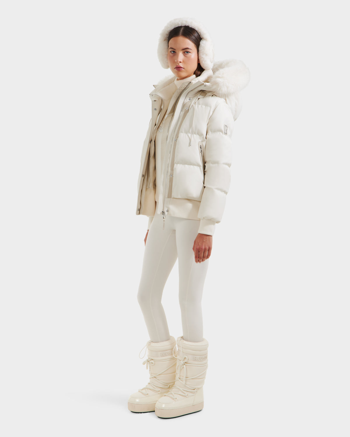 A woman wears the Rudsak CIEYNA - 8125579 Vanilla, an all-white down-filled bomber with a recycled weather-resistant shell, paired with fitted pants and winter boots, standing against a plain white background.