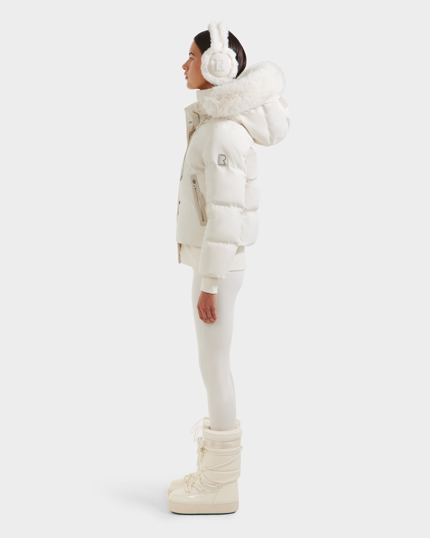 A woman in profile poses before a plain white backdrop, wearing the Rudsak CIEYNA - 8125579 Vanilla recycled shell puffer jacket with fur-trimmed hood, paired with earmuffs, white leggings, and tall snow boots.