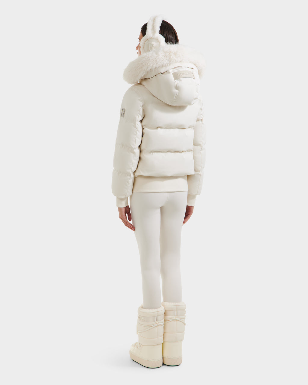 A person faces away, wearing the Rudsak CIEYNA - 8125579 Vanilla all-white winter bomber with a recycled weather-resistant shell, paired with earmuffs, leggings, and insulated boots against a plain white background.