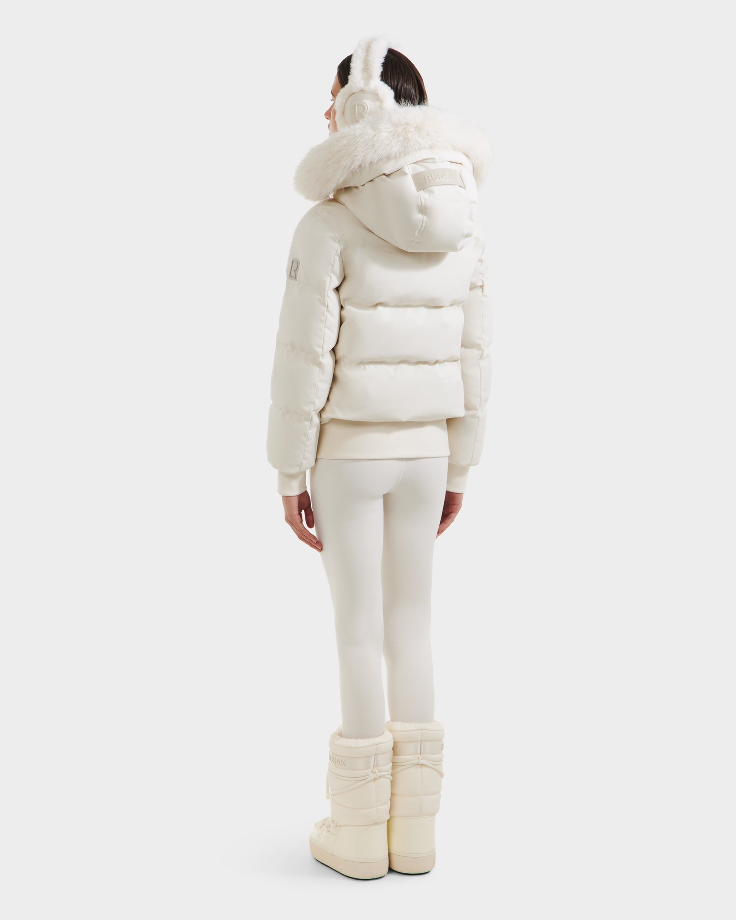 A person faces away, wearing the Rudsak CIEYNA - 8125579 Vanilla all-white winter bomber with a recycled weather-resistant shell, paired with earmuffs, leggings, and insulated boots against a plain white background.