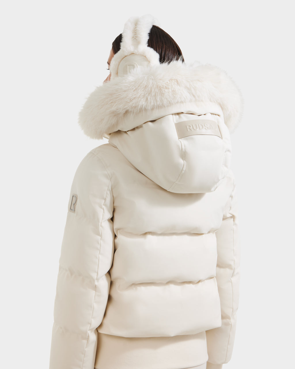 A person seen from the back wears the Rudsak CIEYNA - 8125579 Vanilla cream winter bomber jacket with a large faux fur-trimmed hood and matching earmuffs, set against a plain white background.