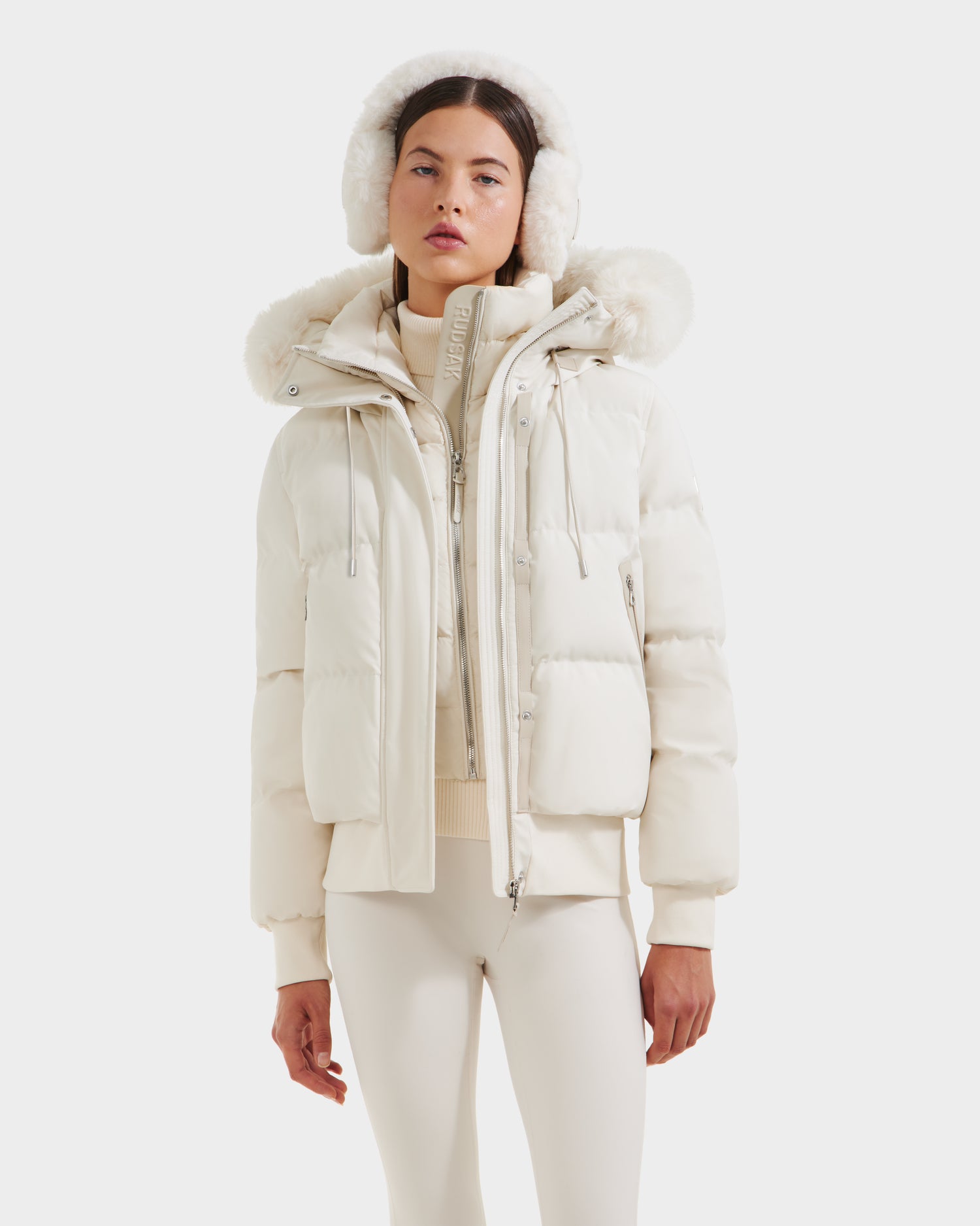 A woman faces forward in the Rudsak CIEYNA - 8125579 Vanilla bomber jacket, featuring a recycled, weather-resistant shell, paired with earmuffs and fitted pants against a plain white background.