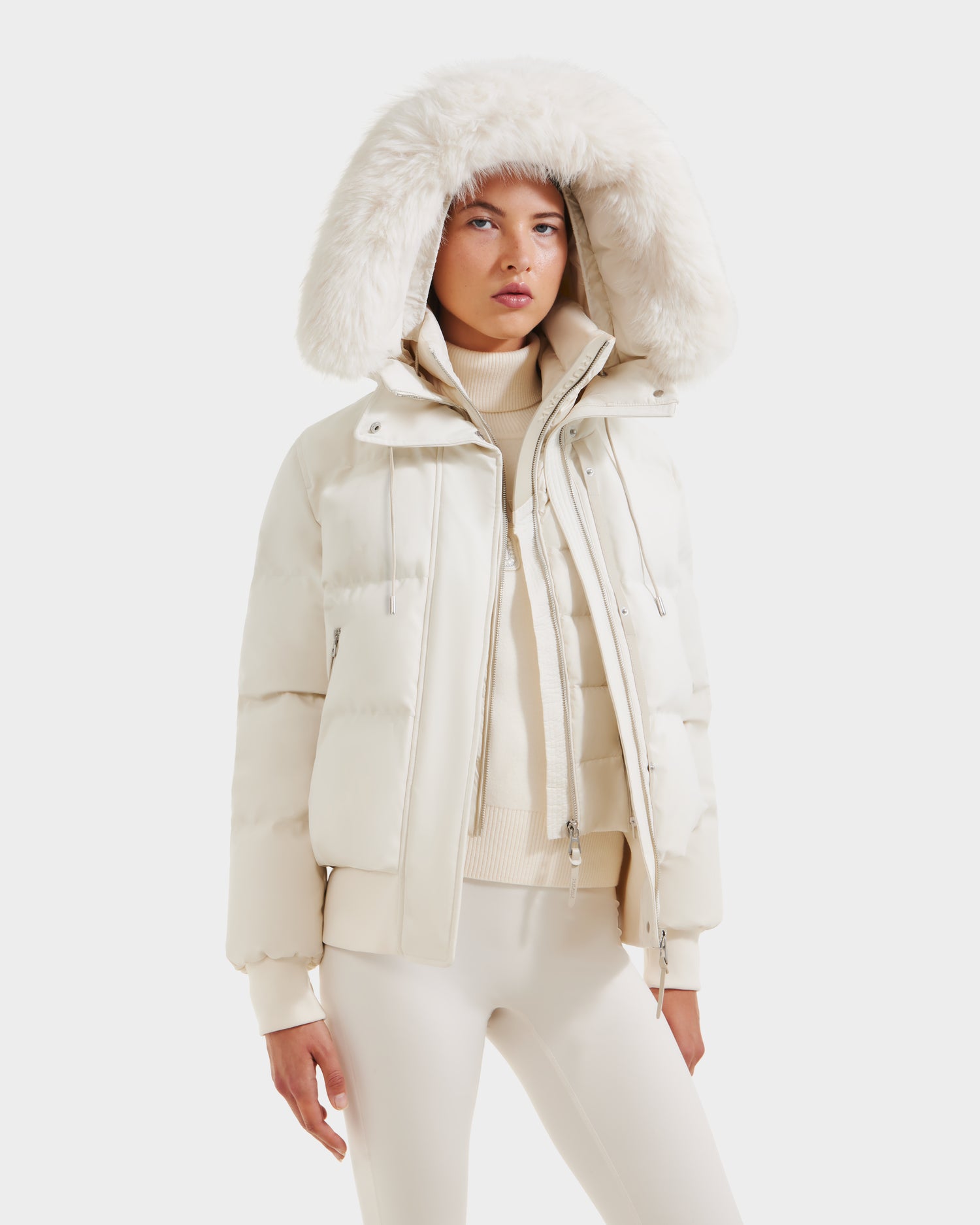 A person wears the Rudsak CIEYNA - 8125579 Vanilla, a white down-filled bomber with a fluffy hood, paired with a cream turtleneck and white pants, standing against a plain white background.