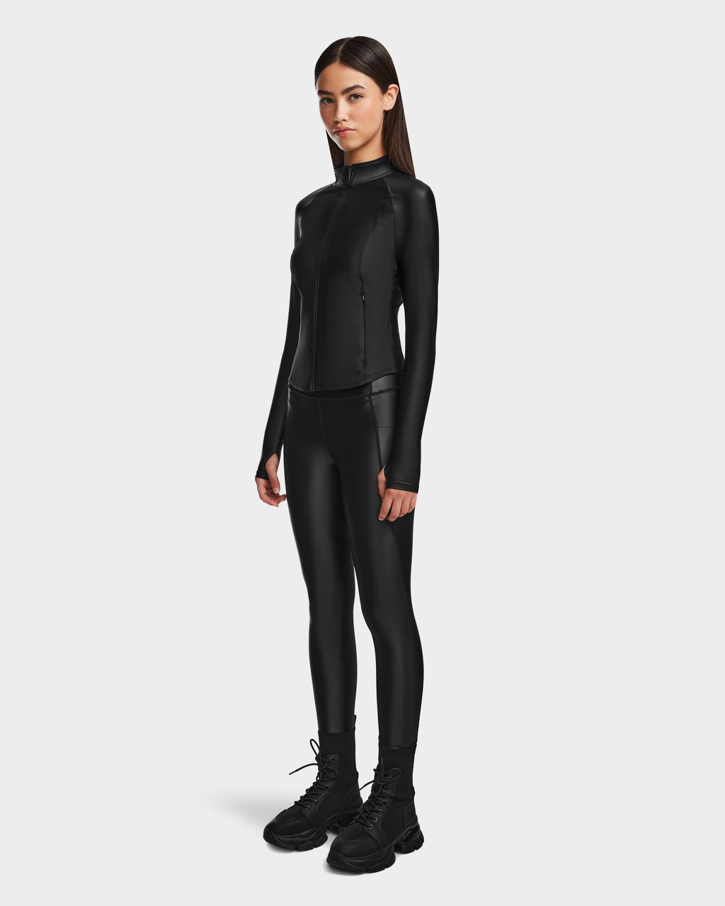 A woman faces forward against a white background, wearing Rudsaks CLARE - 8524519 Black zip-up jacket and womens shiny stretch leggings, paired with chunky black boots.