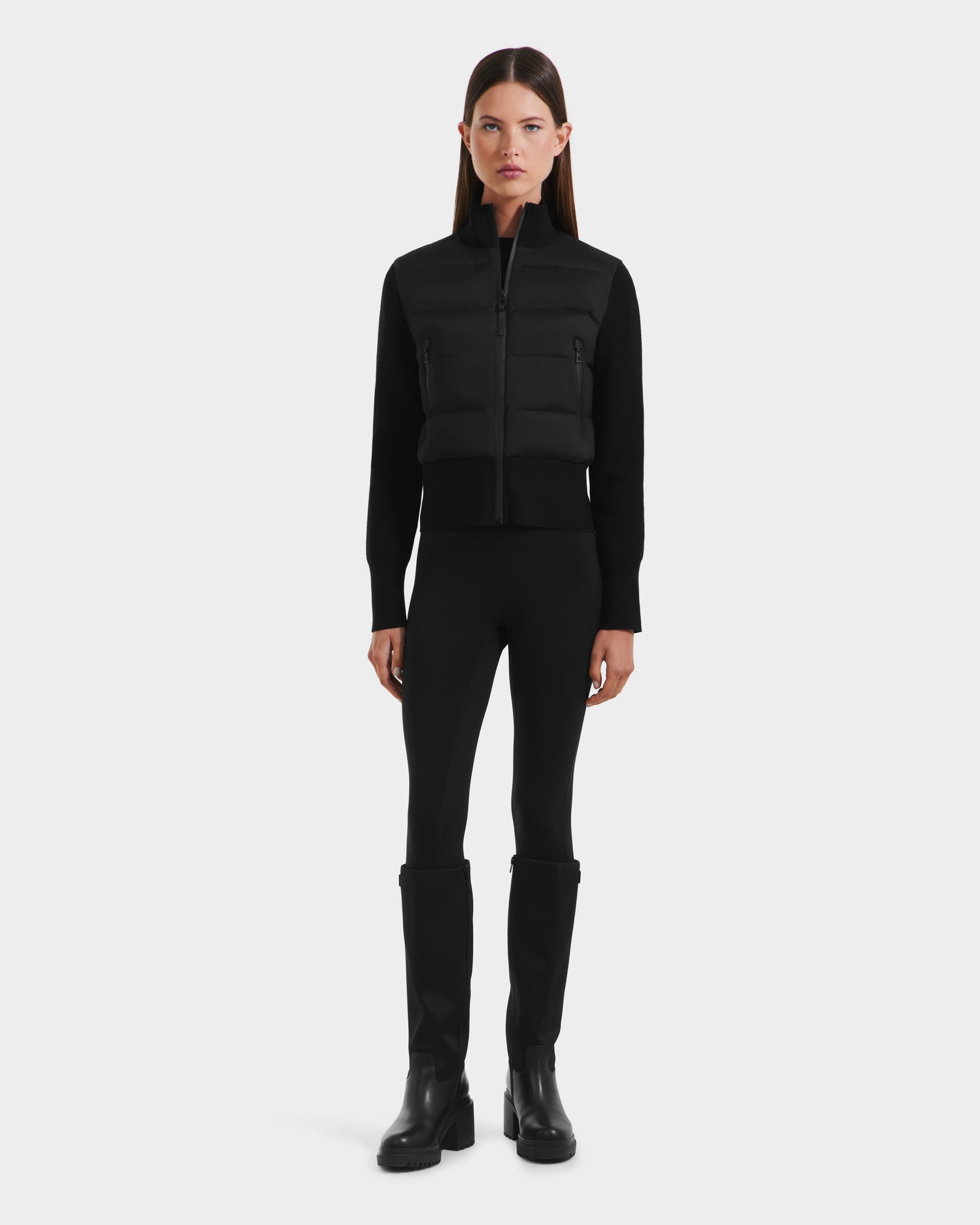 A woman faces forward wearing the Rudsak CLAREMONT - 8125502 Black women’s hybrid down jacket, paired with black fitted pants and black knee-high boots, set against a plain white backdrop.