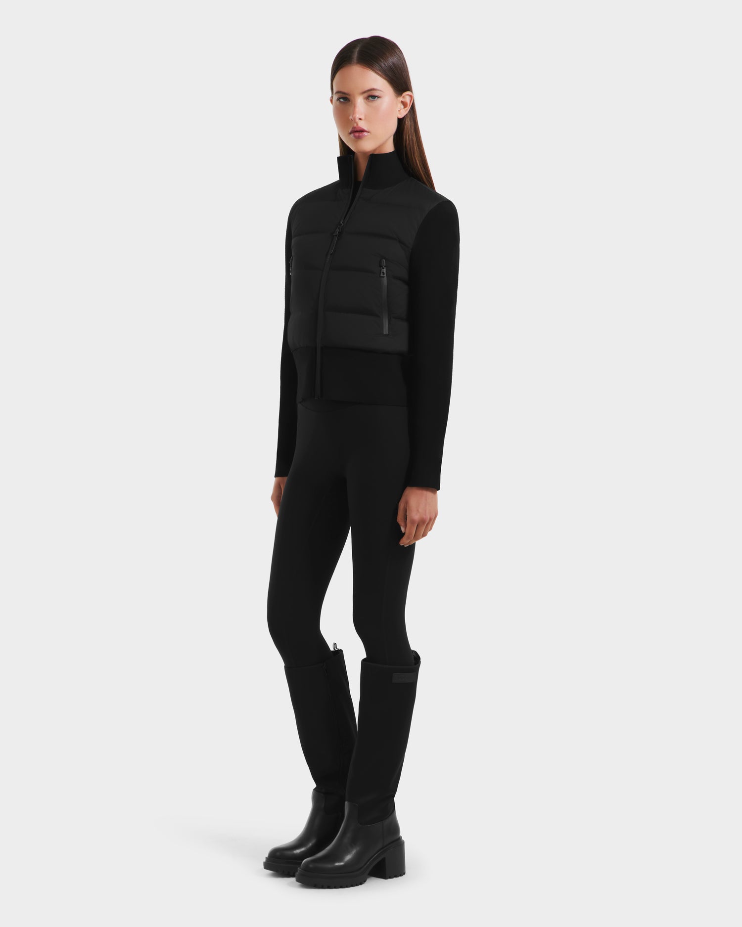 A woman stands facing forward against a plain white background, wearing the Rudsak CLAREMONT - 8125502 Black lightweight warmth jacket over a black long-sleeve top, black leggings, and tall black boots.