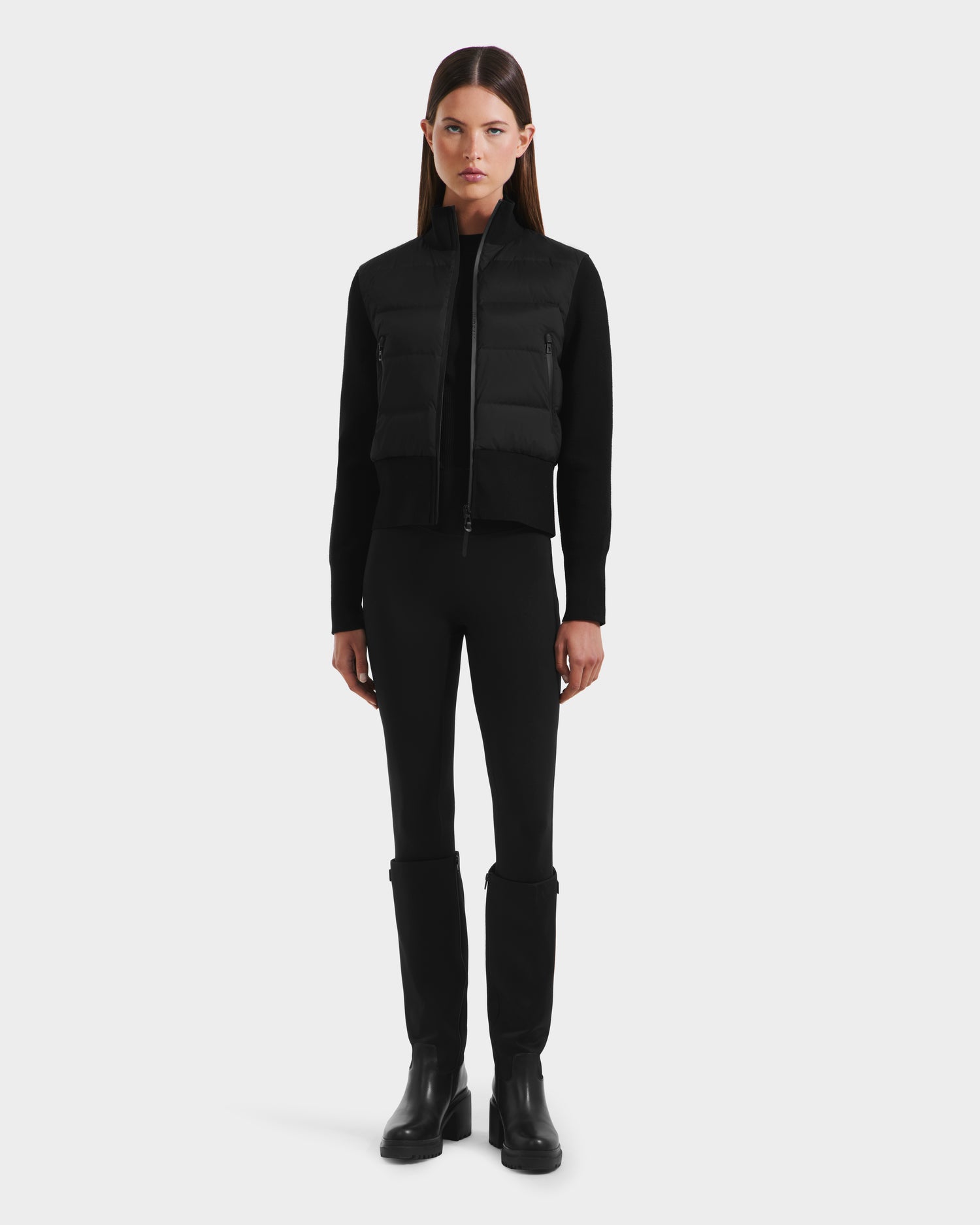 A woman with straight brown hair stands facing forward, wearing the CLAREMONT - 8125502 Black women’s hybrid down jacket by Rudsak, paired with black pants and boots against a plain light background.