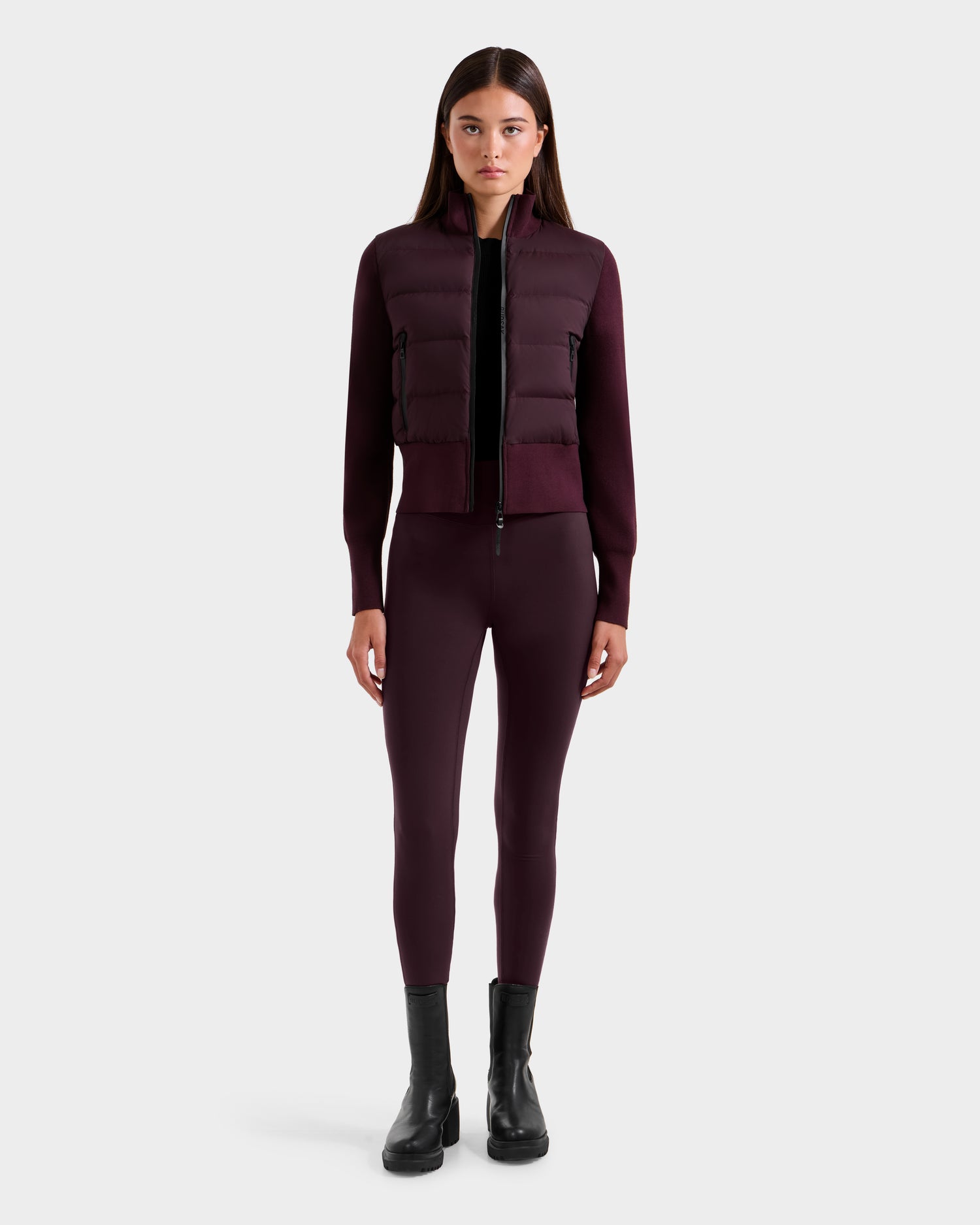 A woman stands facing forward, wearing the Rudsak CLAREMONT - 8125502 Bordeaux women’s hybrid down jacket for lightweight warmth with matching burgundy leggings and black ankle boots against a plain light gray background.