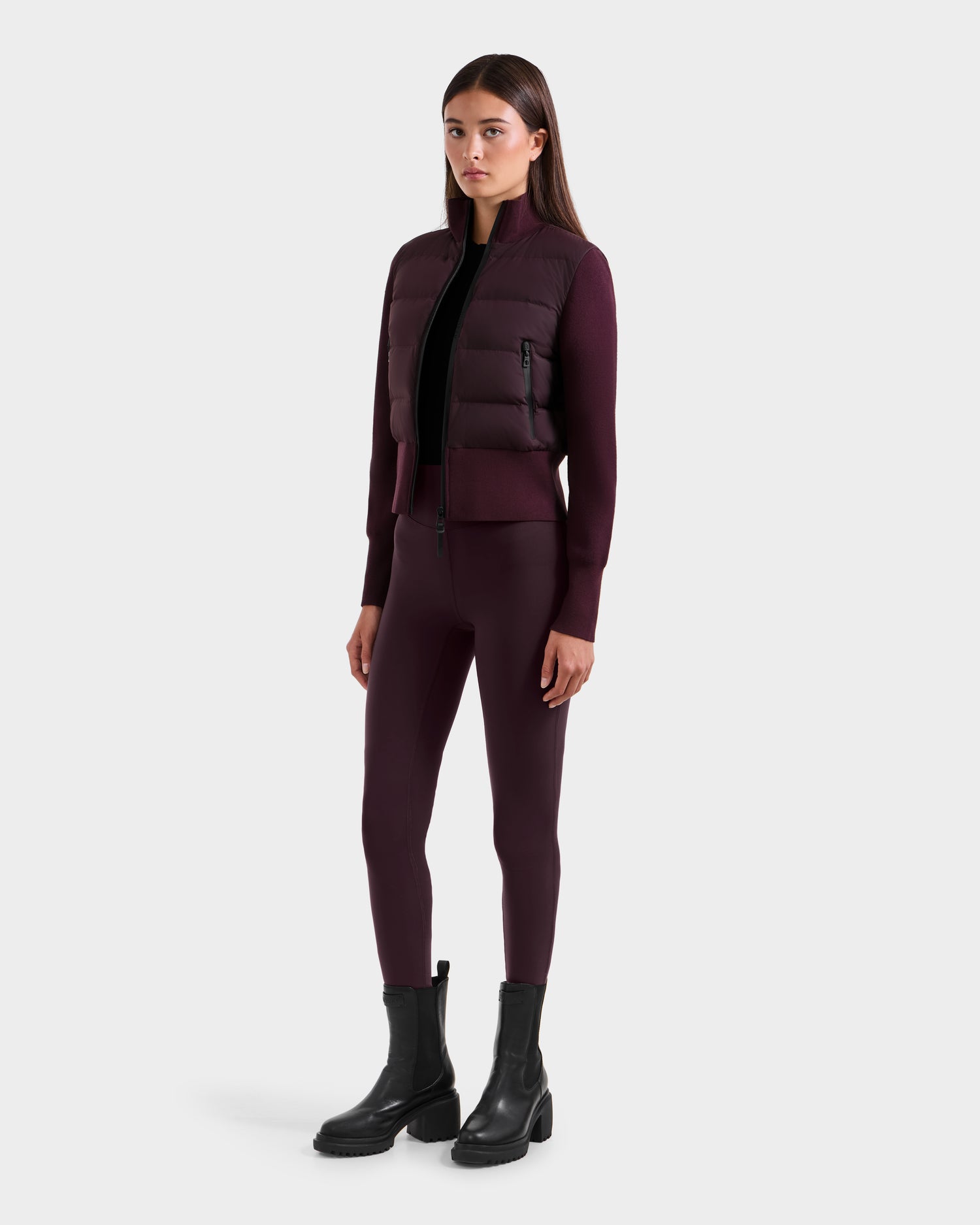 A woman faces forward, wearing the Rudsak CLAREMONT - 8125502 Bordeaux women's hybrid down jacket for lightweight warmth, paired with matching burgundy leggings, a black top, and black ankle boots against a plain light gray background.