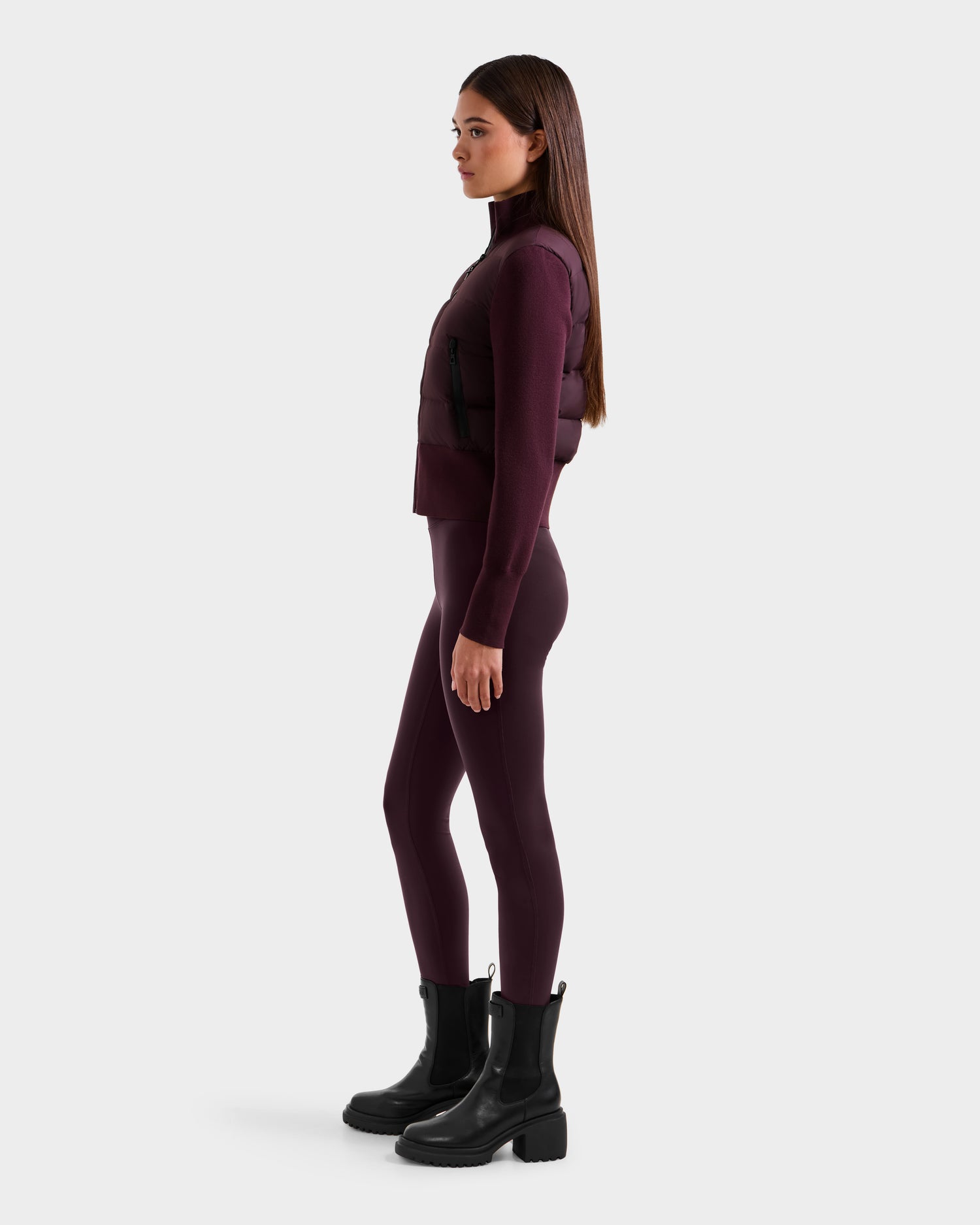 A woman with straight brown hair is in profile, wearing a Rudsak CLAREMONT - 8125502 Bordeaux sleeveless puffer vest, matching leggings, a long-sleeve top, and black ankle boots against a plain white background.