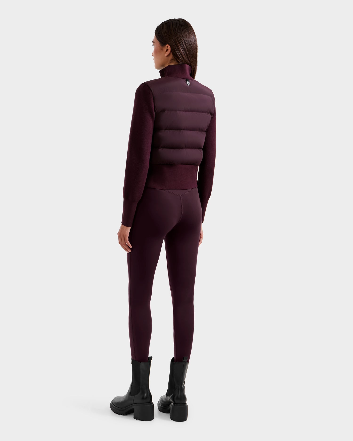 A woman with straight, dark hair stands facing away in the Rudsak CLAREMONT - 8125502 Bordeaux women’s hybrid down jacket, paired with matching leggings and black boots, highlighting its lightweight warmth against a simple background.