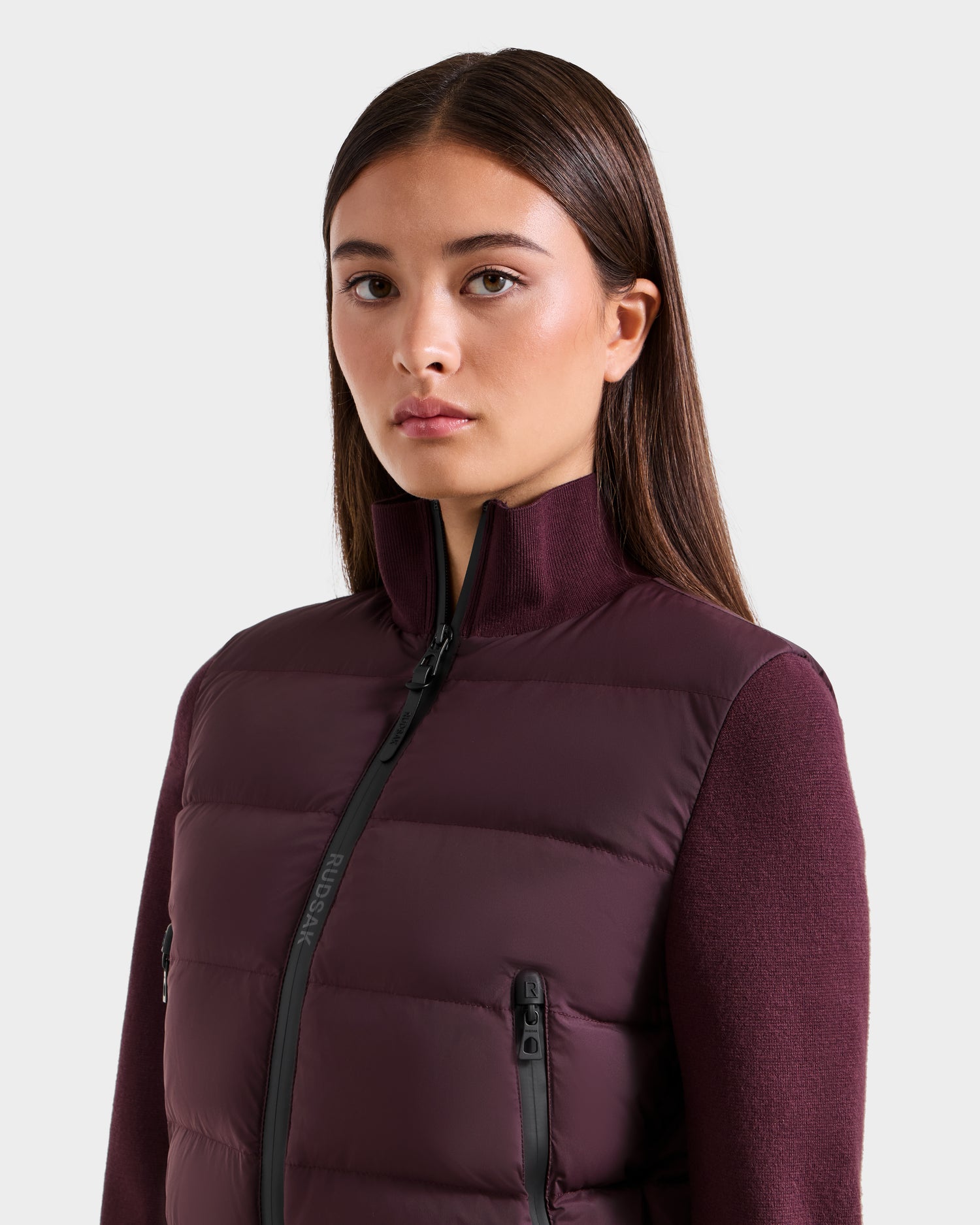 A woman with straight brown hair wears the Rudsak CLAREMONT - 8125502 Bordeaux hybrid down jacket with a high collar and zippered pockets, looking forward against a plain light background—ideal for lightweight warmth.