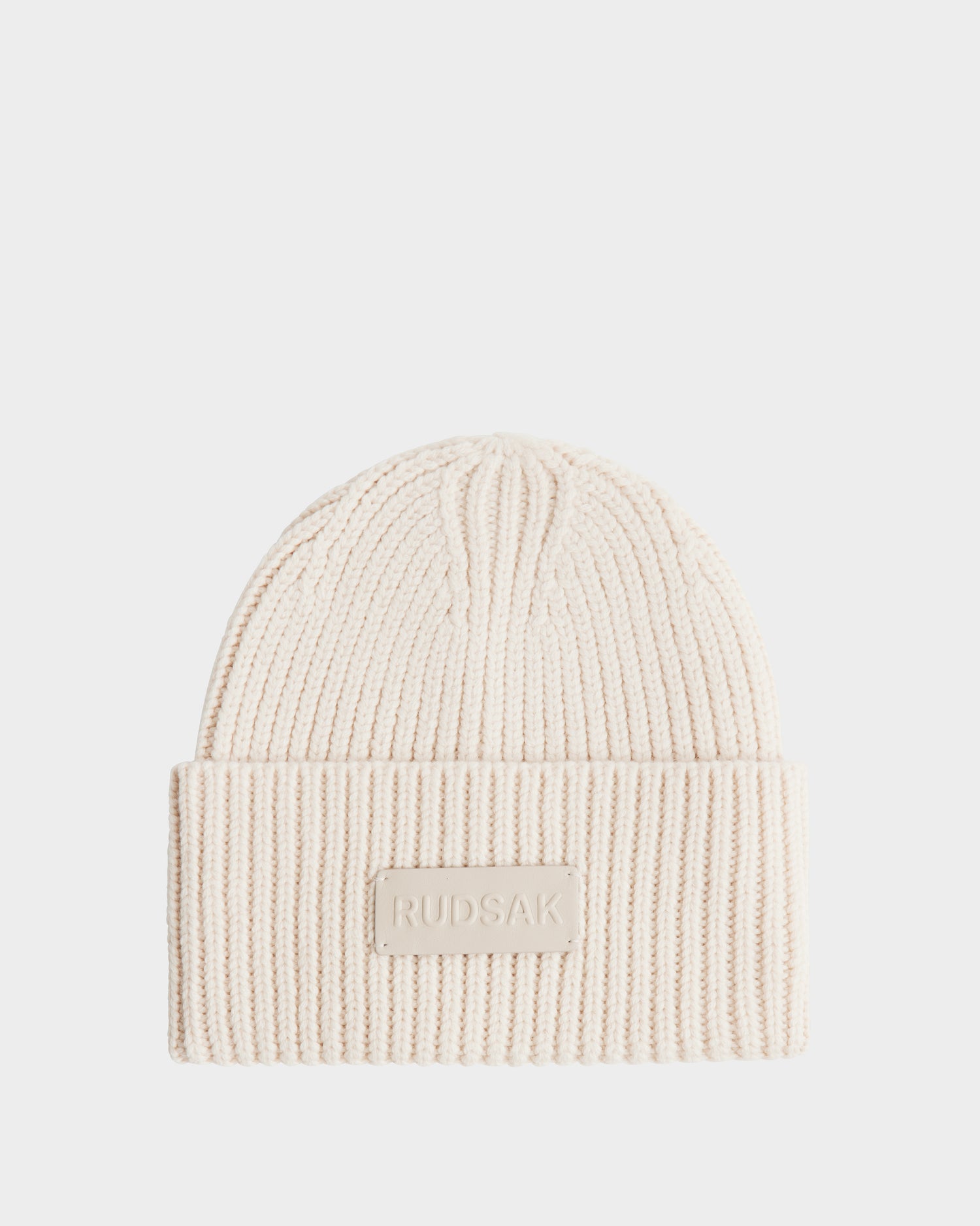 The COLIN - 9424518 Vanilla by Rudsak is a cream ribbed unisex knit beanie with a folded brim and a rectangular patch featuring the Rudsak name in raised letters on the front.