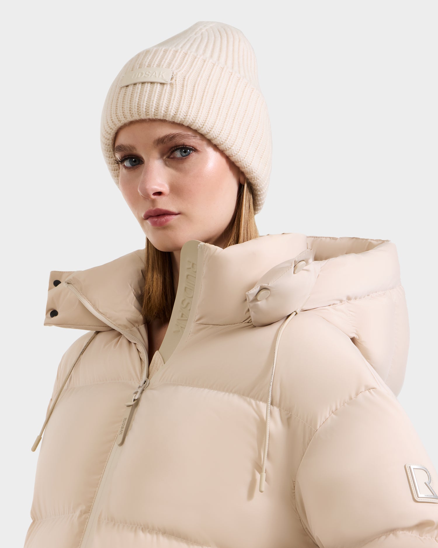 A woman in the Rudsak COLIN - 9424518 Vanilla puffer jacket and a matching beanie, both with subtle branding, looks ahead with a neutral expression. The high-collar jacket and accessories are set against a plain white background.
