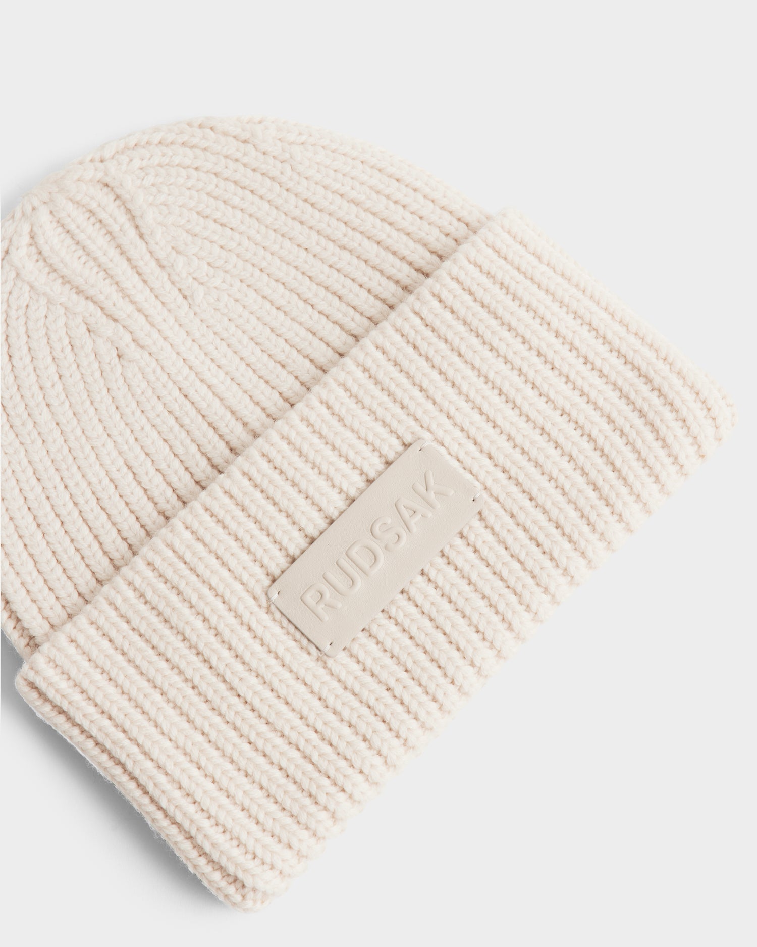 The COLIN - 9424518 Vanilla is a cream ribbed knit unisex beanie with a folded brim and a rectangular patch displaying the Rudsak brand name in raised letters.