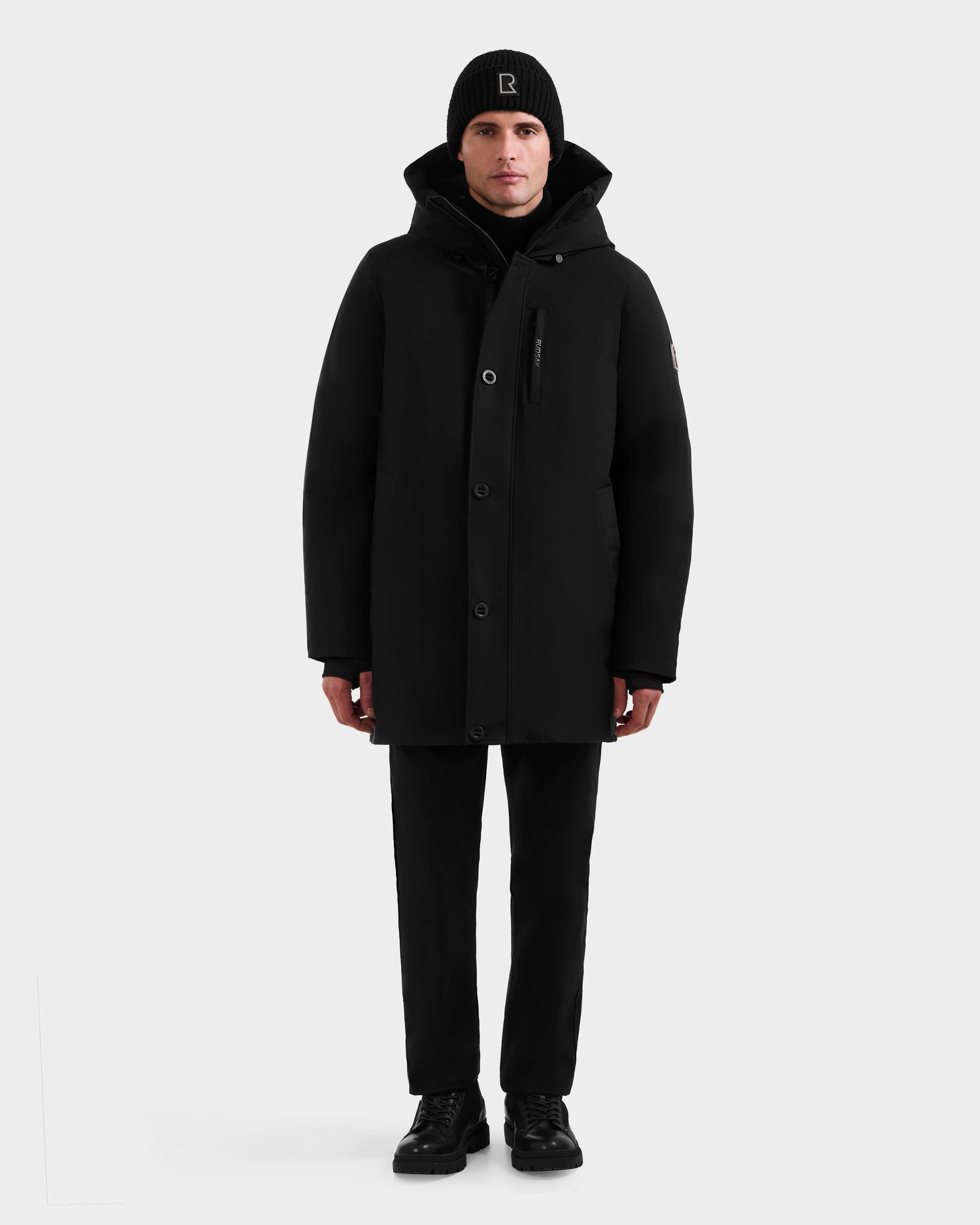A person stands facing forward, wearing the Rudsak COREY - M125512 Black men’s down parka with RDS-certified down, black pants, black shoes, and a black beanie with a white logo against a plain light background.