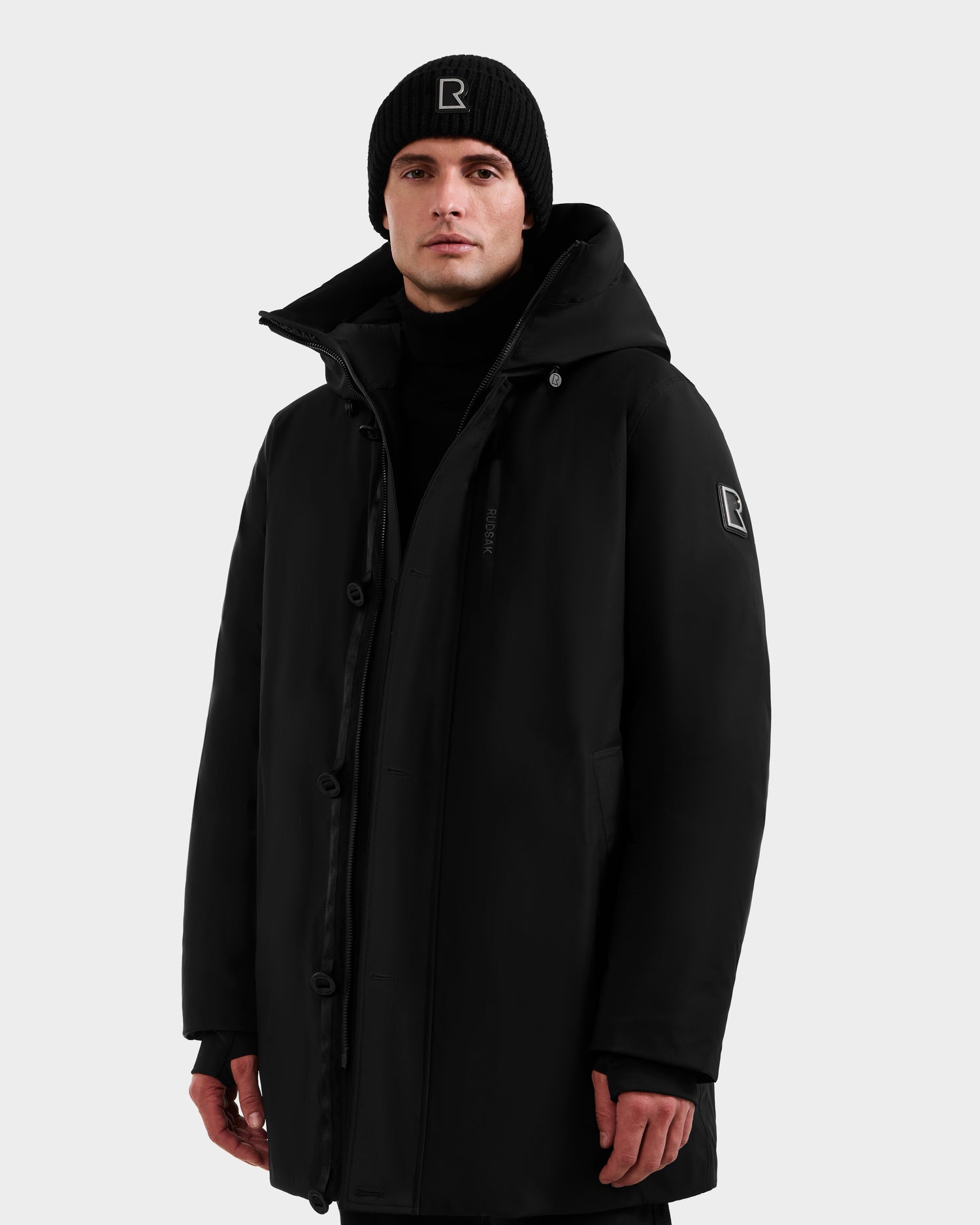 A person wearing the Rudsak COREY - M125512 Black men’s down parka with a hood, RDS-certified fill, and sleeve logo patch, plus a black beanie with a rectangular logo, stands against a plain white background.