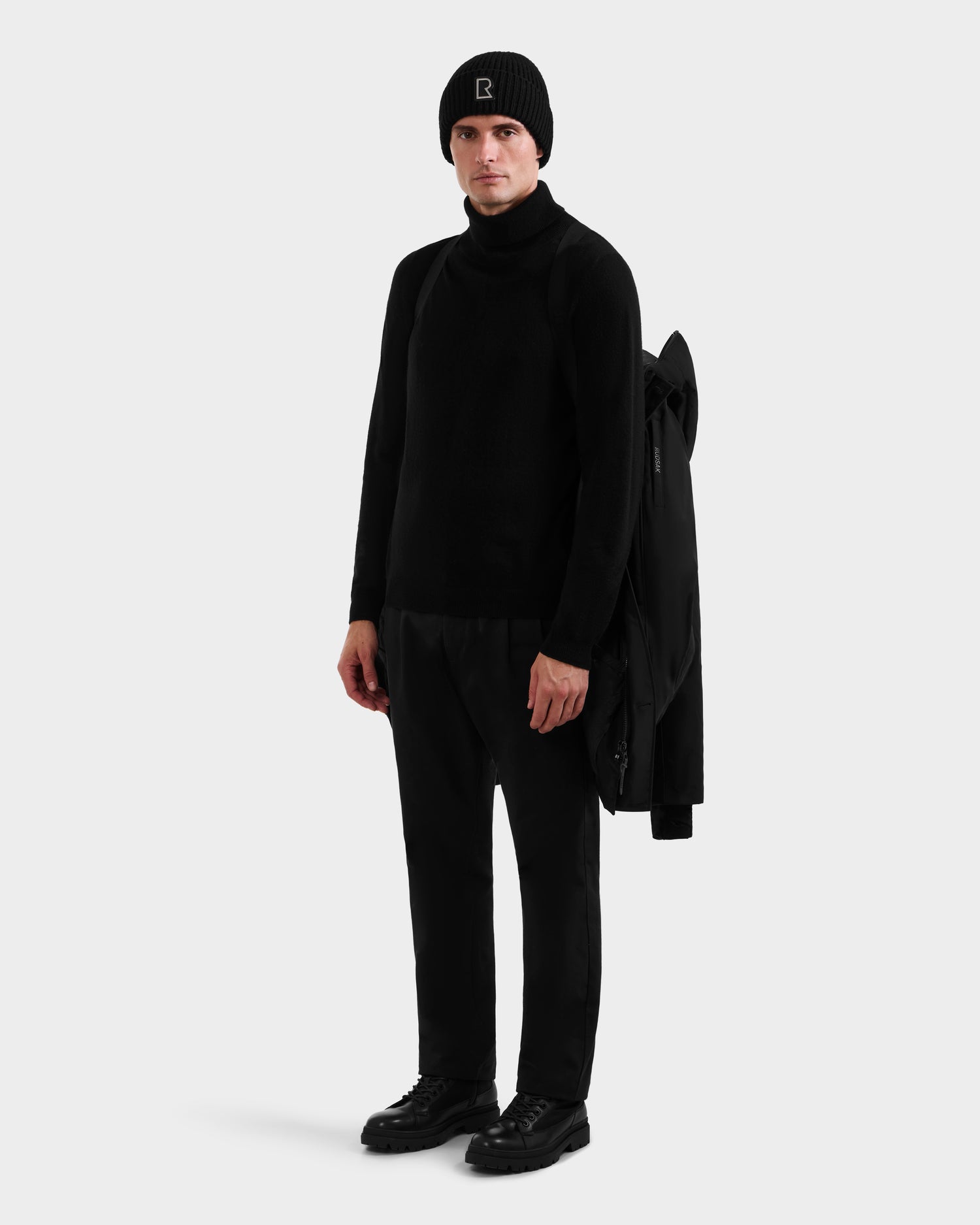 A person faces forward against a plain light background, dressed in all black with Rudsak’s COREY - M125512 Black men’s down parka draped over one shoulder.