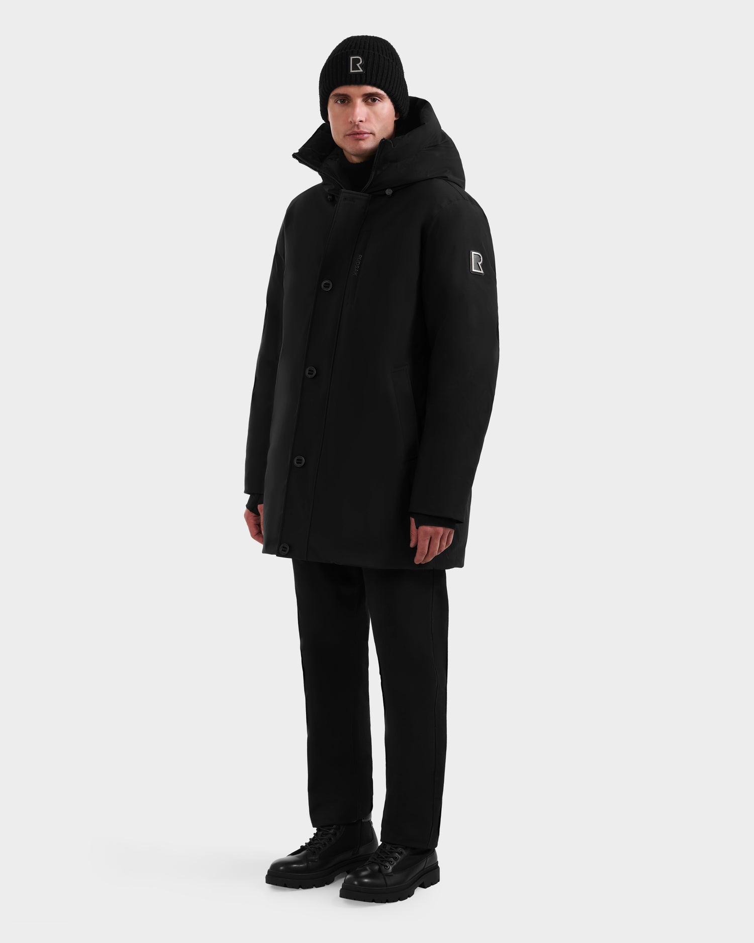 A person in the Rudsak COREY - M125512 Black parka with RDS-certified down, matching black beanie, pants, and shoes stands against a white background. A small white logo appears on the coat sleeve and beanie.