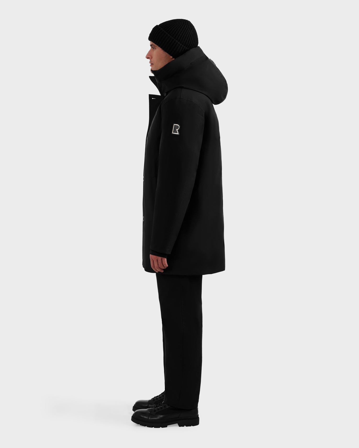 A person stands in profile against a plain background, wearing the Rudsak COREY - M125512 Black RDS-certified down parka with hood, black pants, black shoes, and a black knit beanie. A square patch appears on the upper left sleeve of the coat.