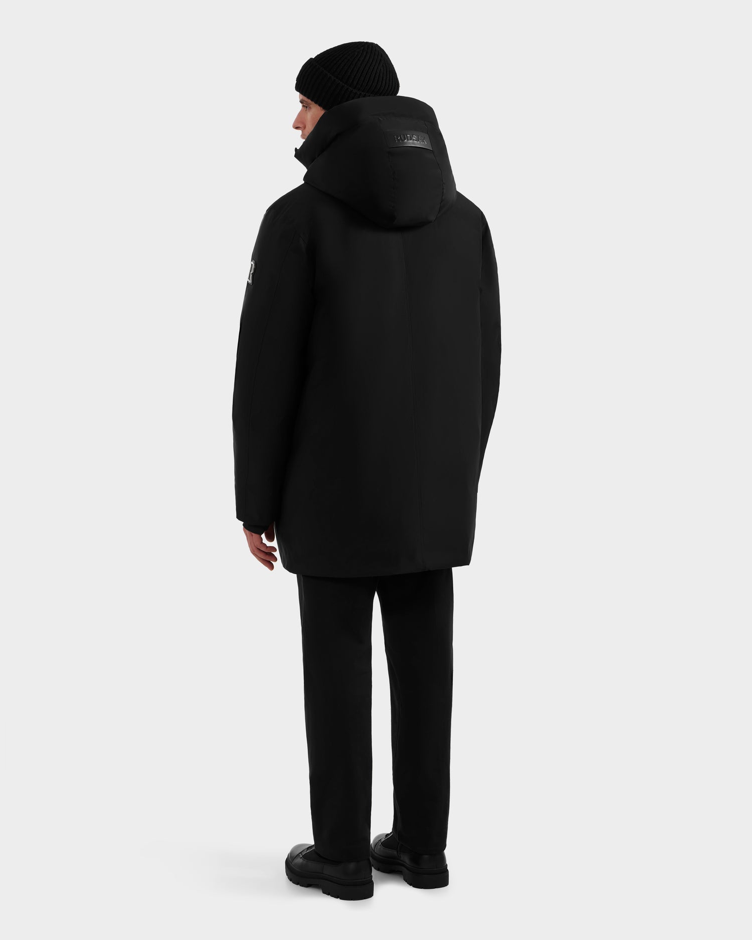 A person stands facing away from the camera, wearing the Rudsak COREY - M125512 Black men's down parka with RDS-certified down, paired with black pants, boots, and a knit beanie against a plain light background.