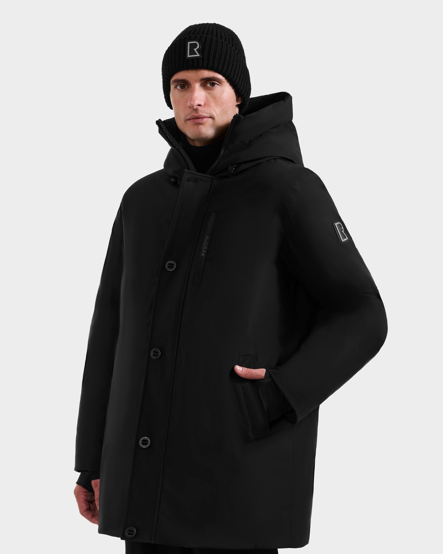 A man in the Rudsak COREY - M125512 Black, a men's down parka with RDS-certified down and zipper-button closure, wears a black beanie and stands against a plain light background with one hand in his pocket.