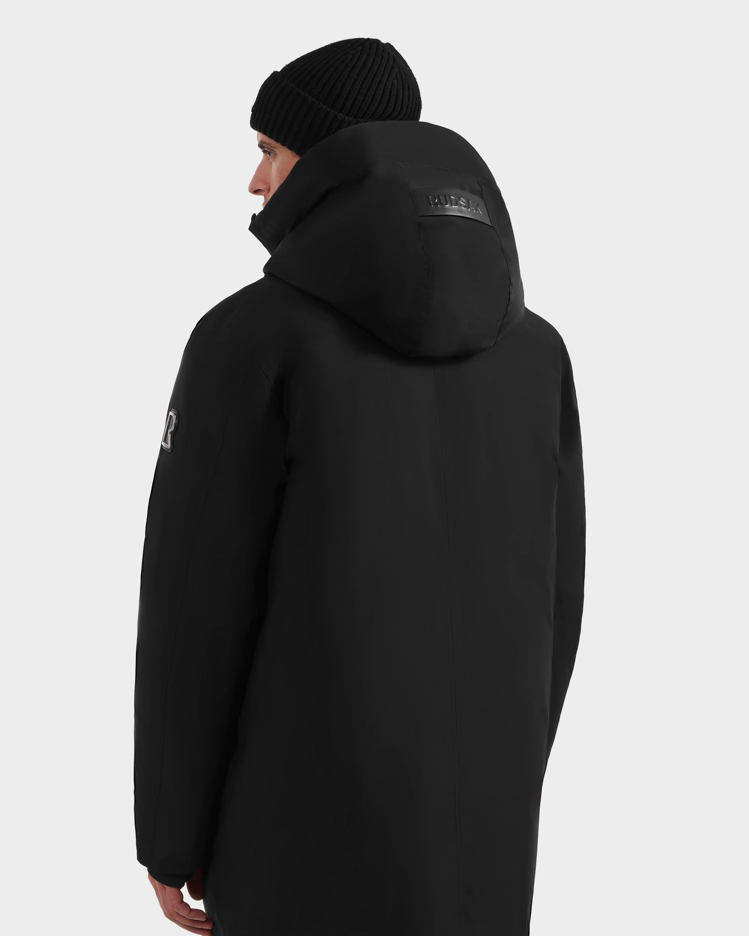A person is seen from behind wearing the Rudsak COREY - M125512 Black men’s down parka and a beanie. The black parka features a patch on the hood and an emblem on the upper left sleeve, set against a plain white background.