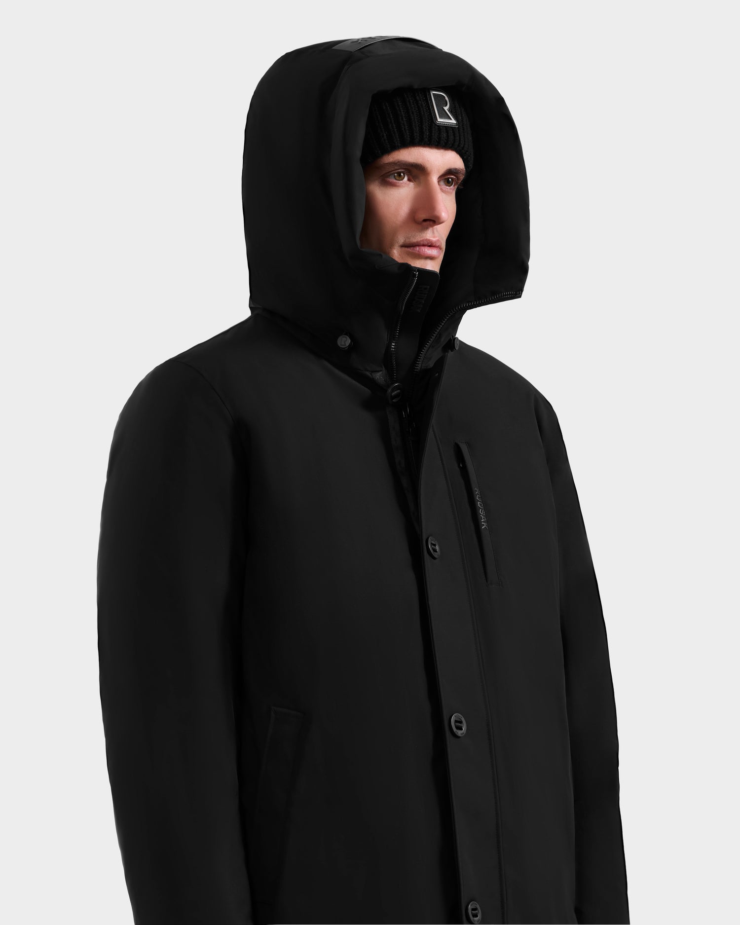 A person wearing the Rudsak COREY - M125512 Black men’s down parka and a black knit beanie stands against a plain light background, looking slightly to the side.