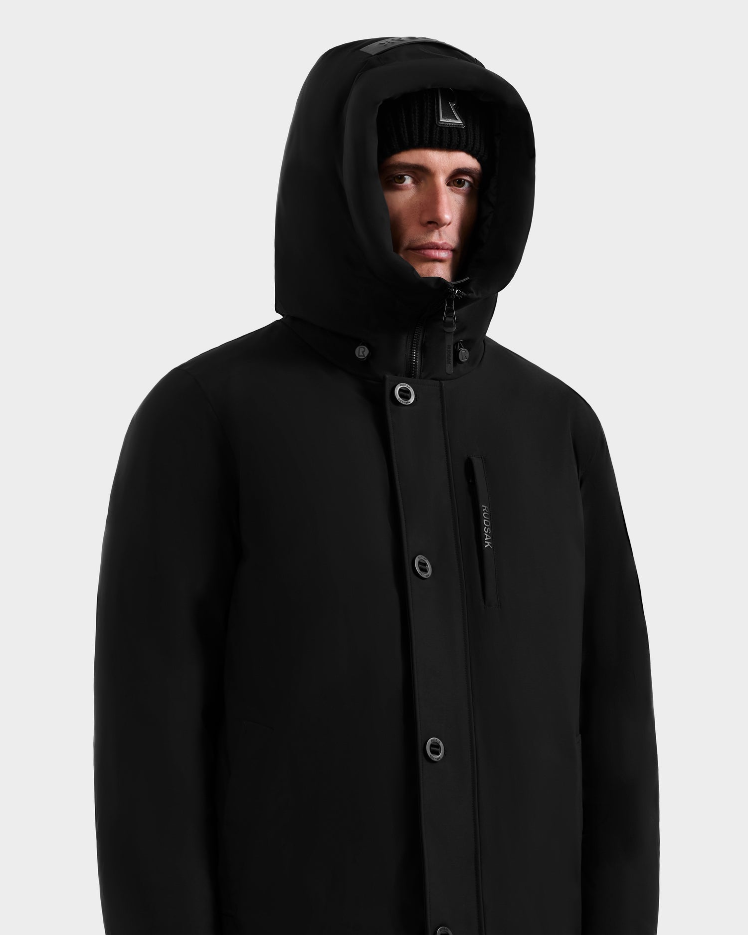 A person wearing the Rudsak COREY - M125512 Black men’s down parka and a black beanie, looking forward with a neutral expression against a plain light gray background.