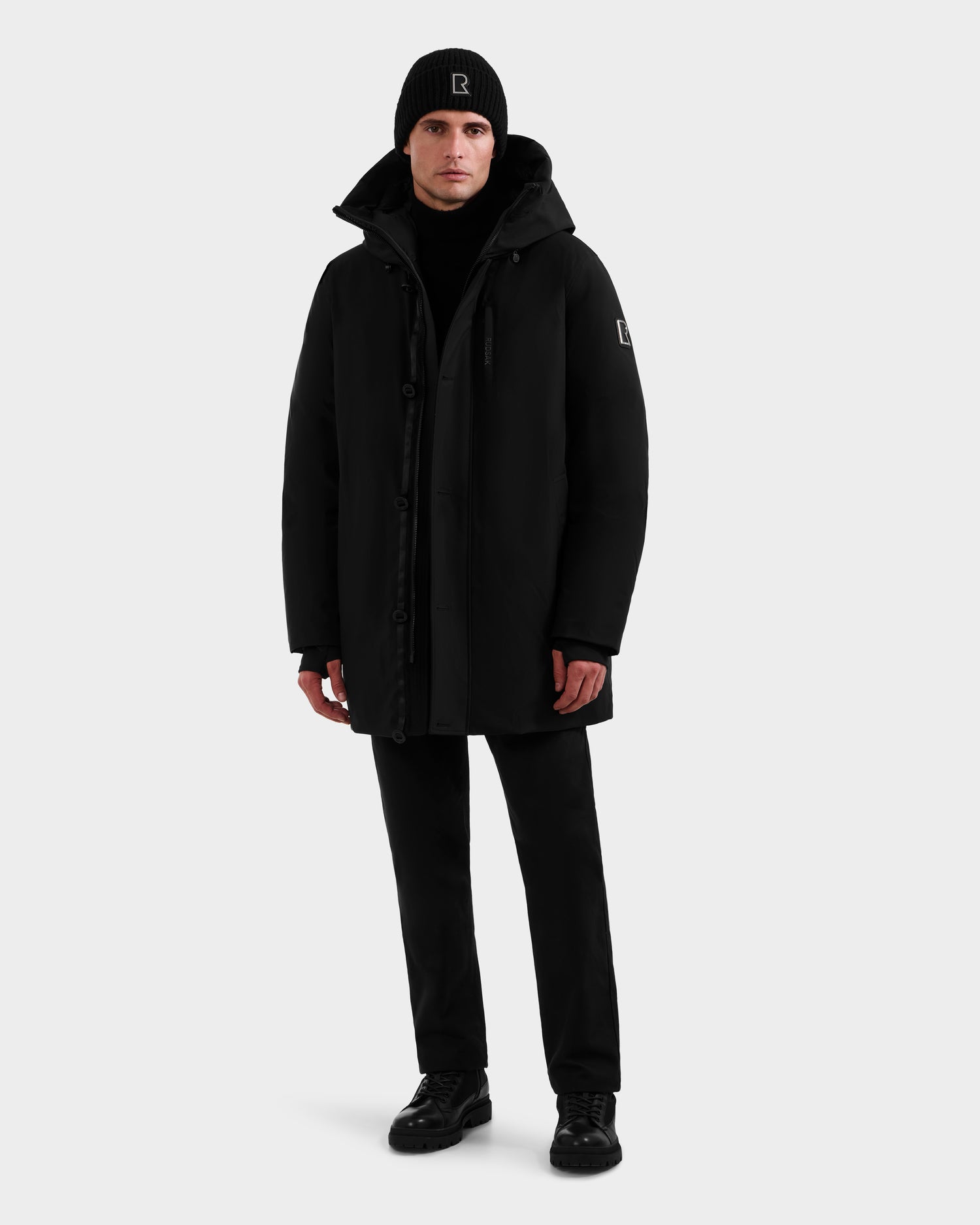 A person faces forward wearing the Rudsak COREY - M125512 Black parka, black pants, a matching beanie, and black shoes against a plain light background.