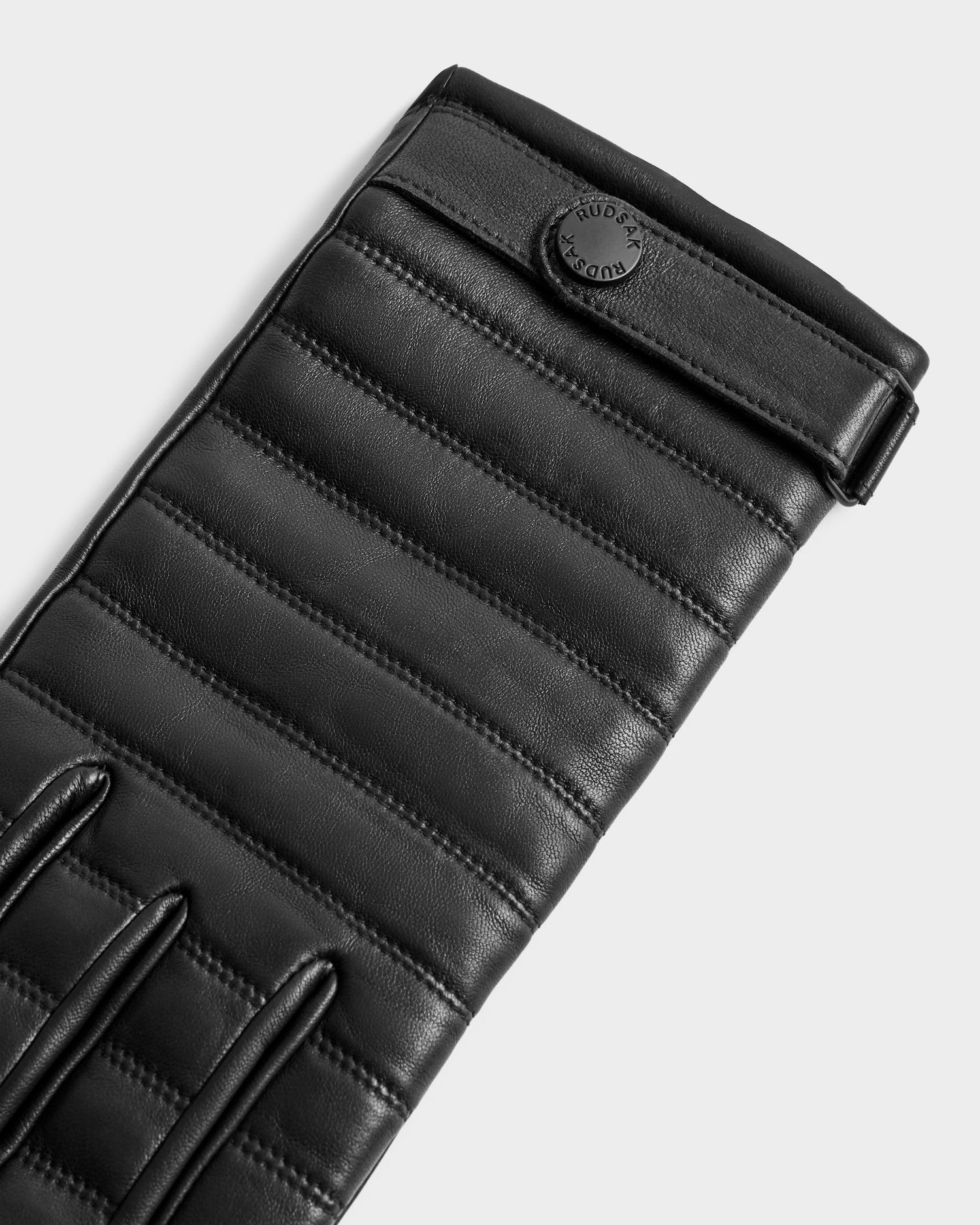 Close-up of Rudsak CORTES - 6425036 Black leather gloves with horizontal padded stitching and a circular embossed button at the wrist. These stylish touchscreen winter gloves stand out against a plain, light background.