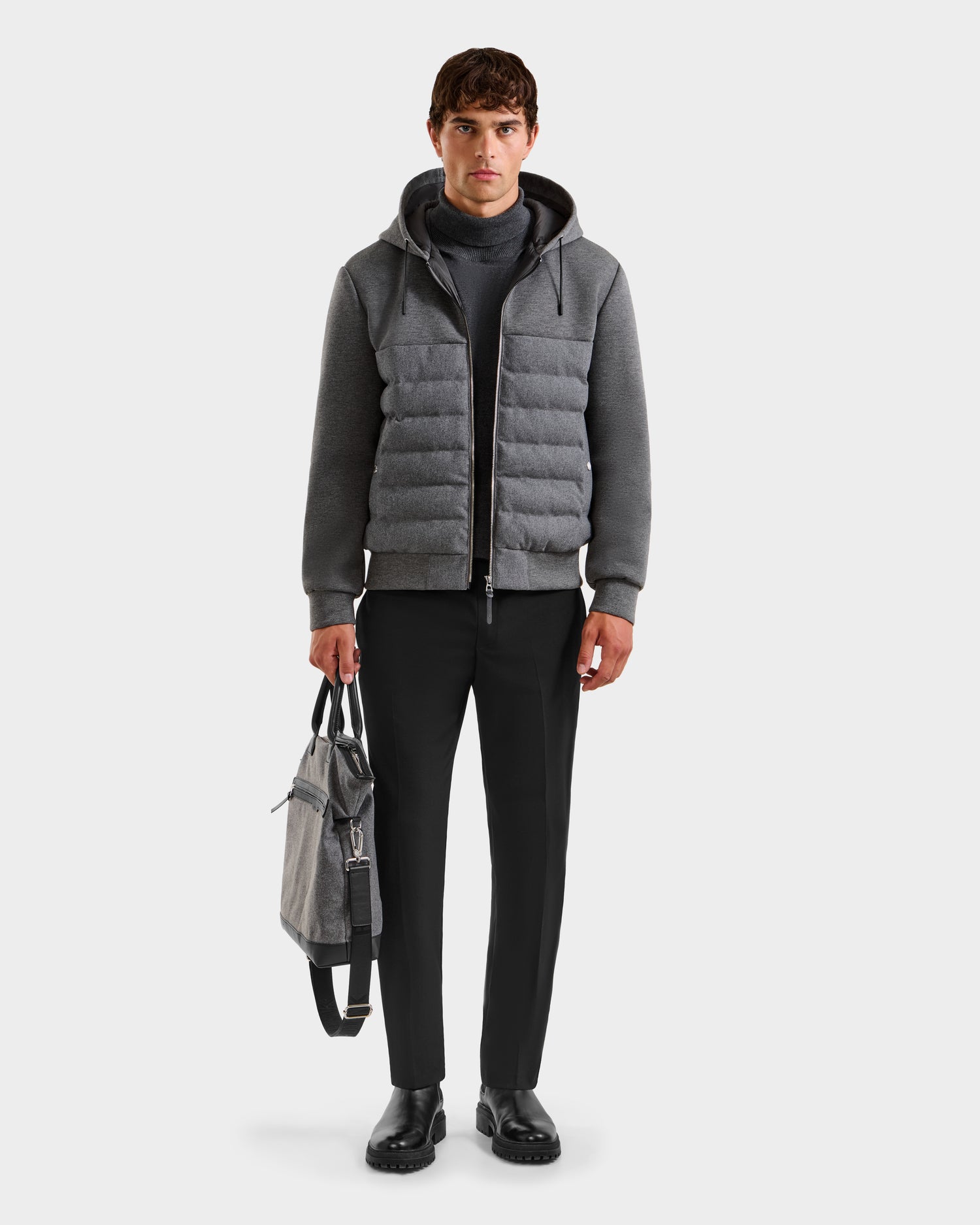 A man faces forward wearing the Rudsak CURTIS W - 6125581 lightweight down jacket in gray, styled with a dark turtleneck, black pants, and shoes. He holds a gray bag in his left hand against a plain white background.