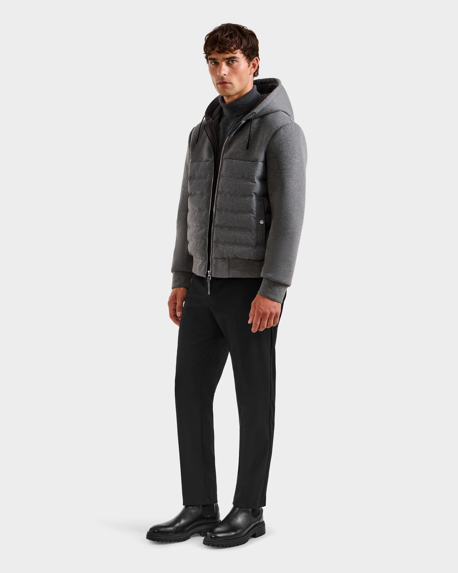 A man stands slightly left, wearing the Rudsak CURTIS W - 6125581 lightweight down jacket in grey with a hood, paired with black pants and boots against a plain white background.