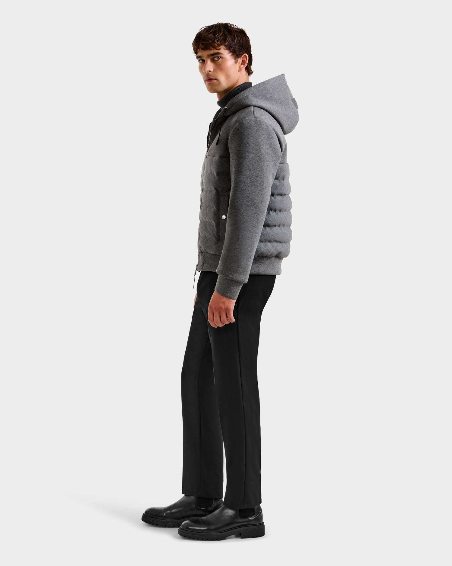 A young man in profile wears the Rudsak CURTIS W - 6125581 Grey lightweight down jacket with a gray hood over a dark shirt, black pants, and black shoes, standing against a plain white background.