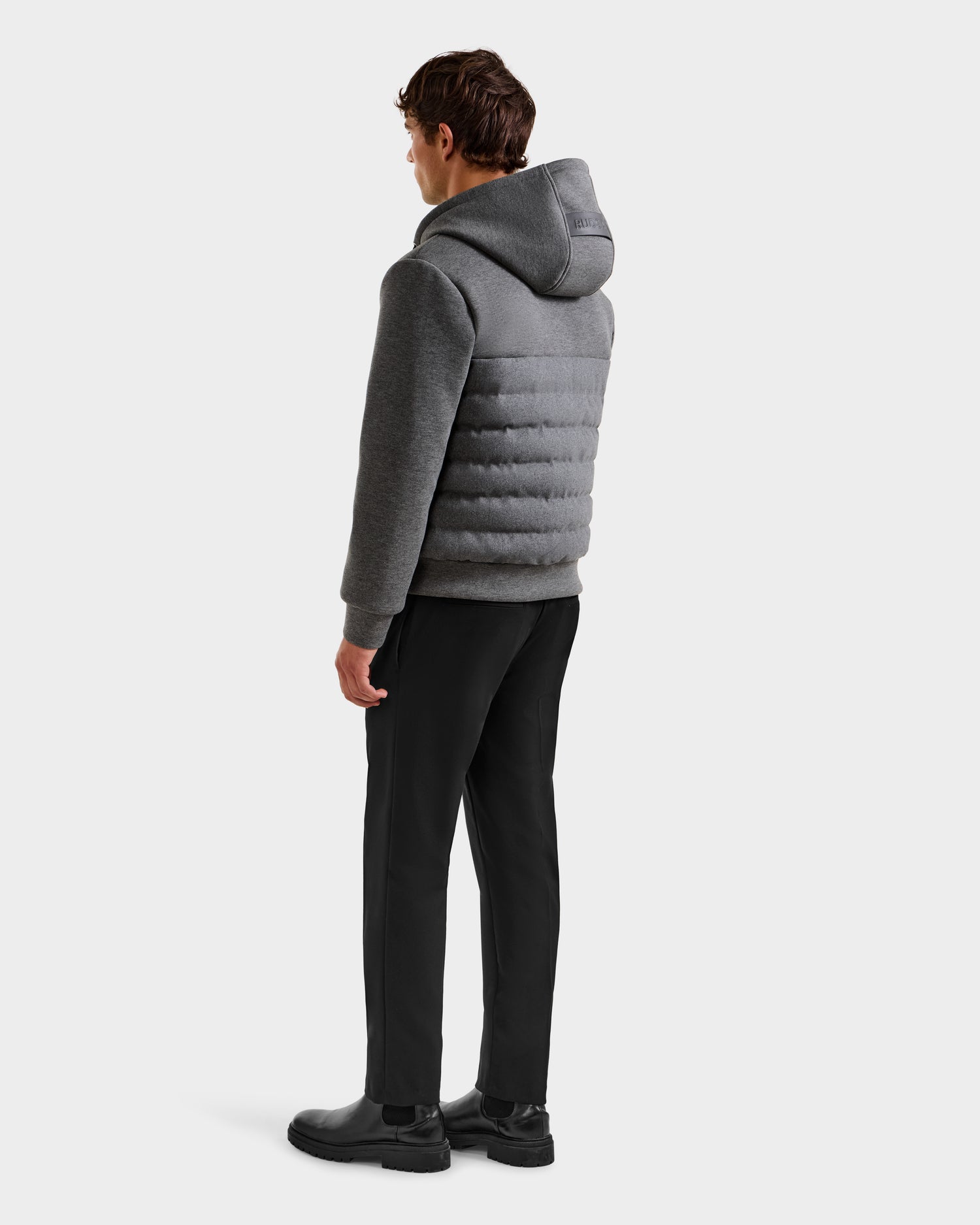 A person stands facing away, wearing the Rudsak CURTIS W - 6125581 Grey men’s lightweight down jacket with a gray hood, paired with black pants and black boots against a plain white background.