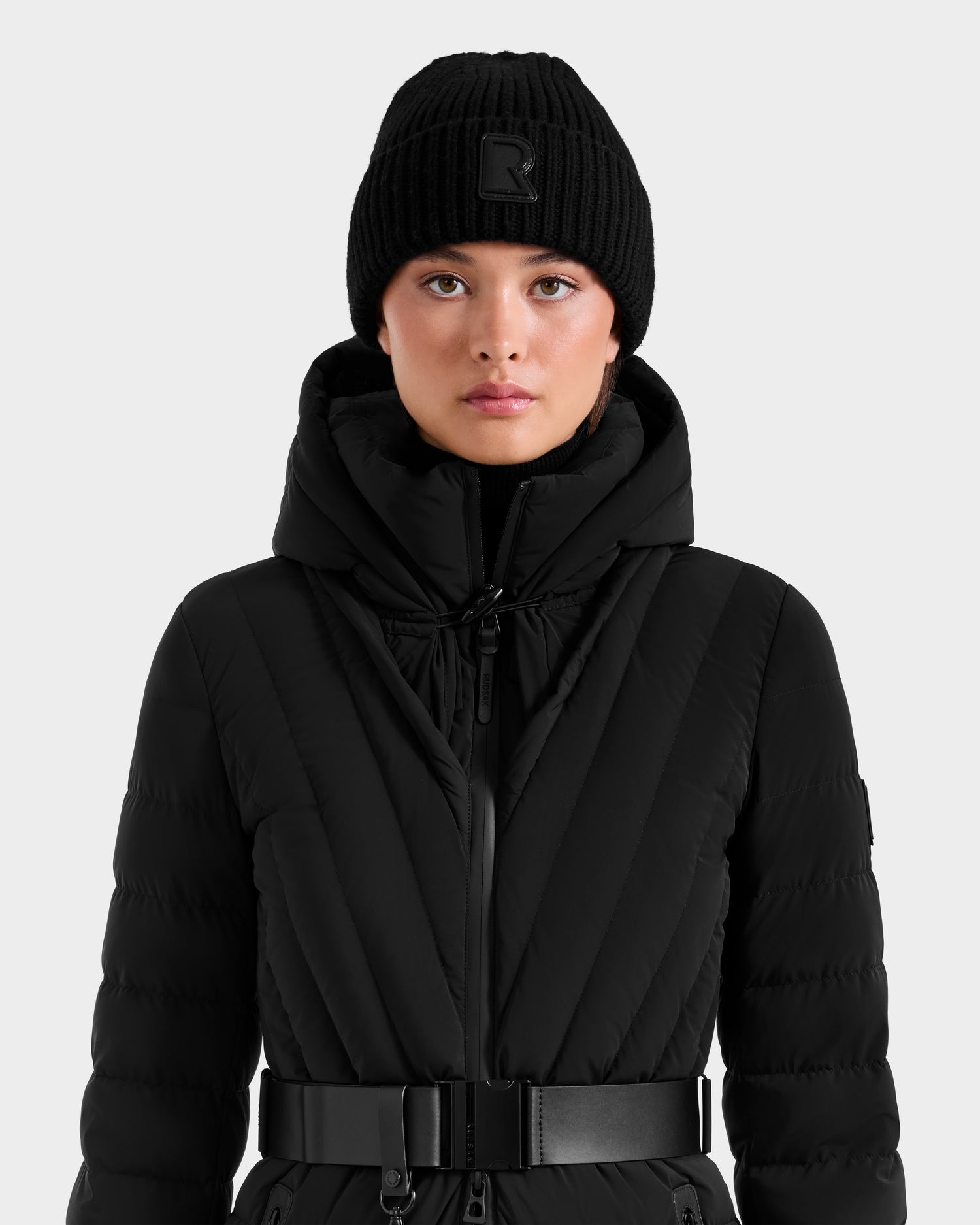 A woman in the Rudsak CYBELLE - 9424504 Black quilted winter coat with belt and a wool blend beanie faces forward against a plain white background, highlighting this stylish outerwear piece.