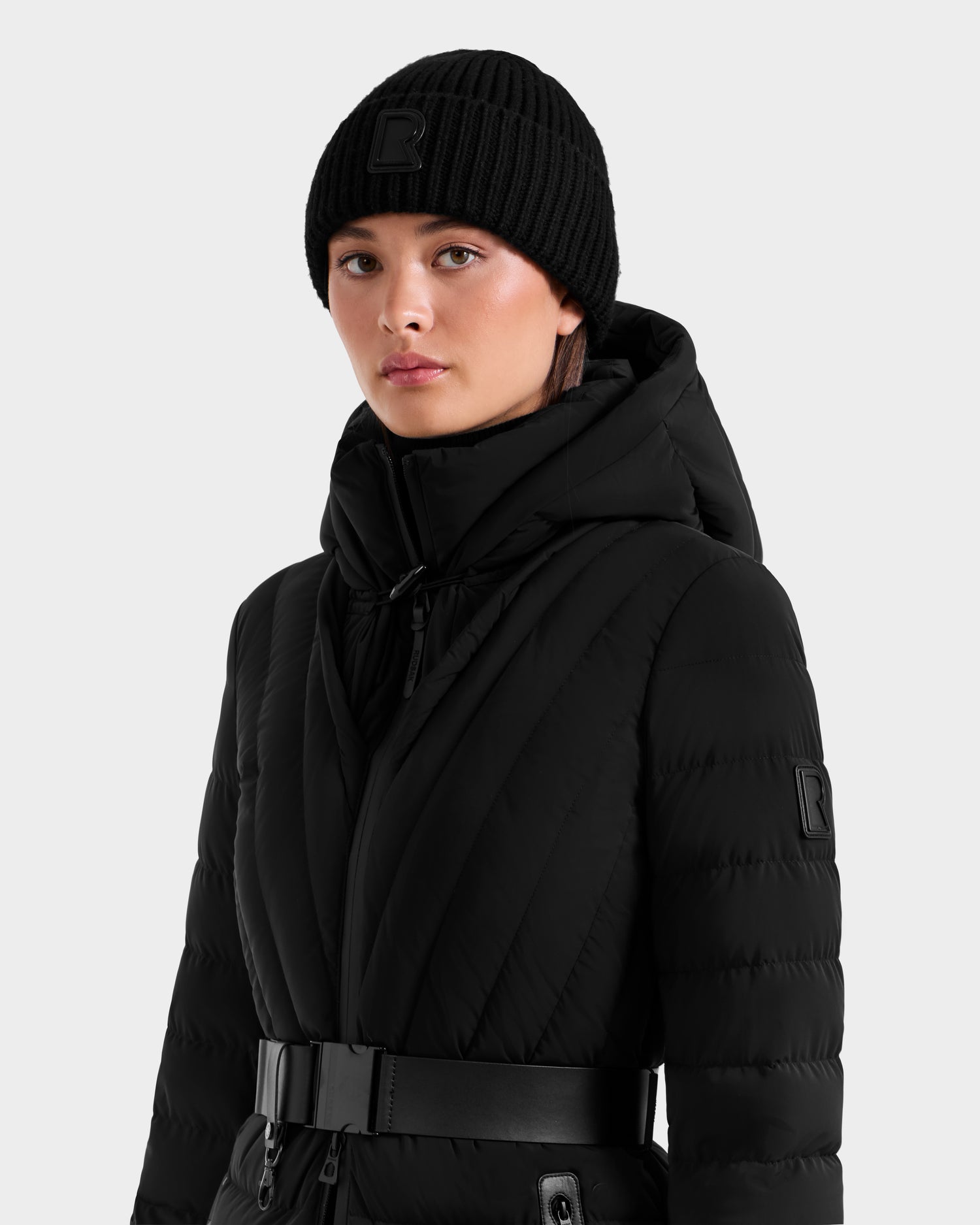 A person wears Rudsak’s CYBELLE - 9424504 Black puffer coat with a wide belt and matching wool blend beanie, both showcasing a raised R logo, against a plain light grey background—a chic winter look.