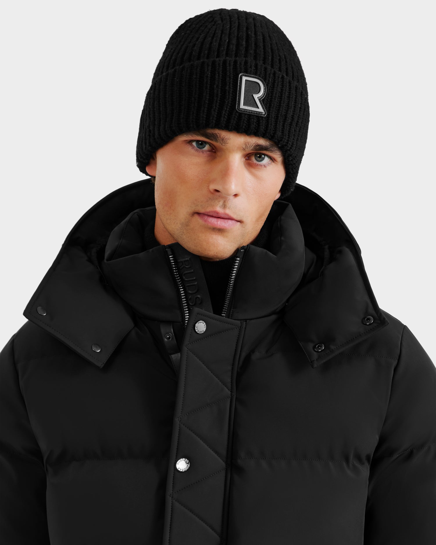 Wearing the Rudsak CYBELLE - 9424504 Black/Silver quilted jacket with large buttons and a high collar, and a black unisex beanie with a white R logo, a person poses against a plain light background.