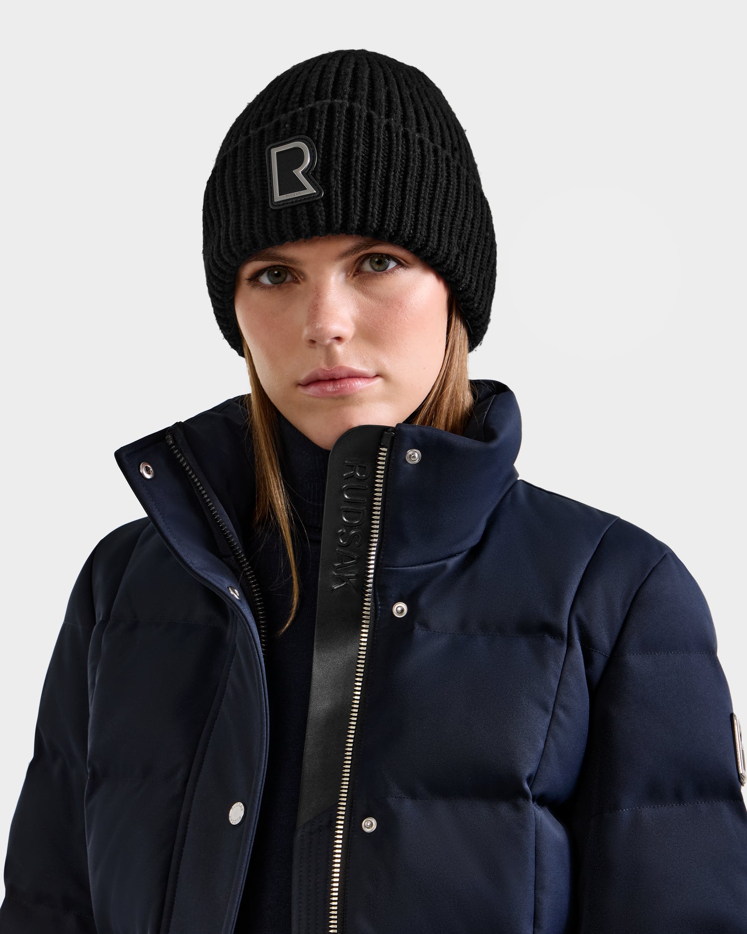 A person wearing Rudsak’s CYBELLE - 9424504 Black/Silver wool-blend beanie with an R logo, styled with a navy puffer jacket and black zip-up layer, stands against a plain light background.