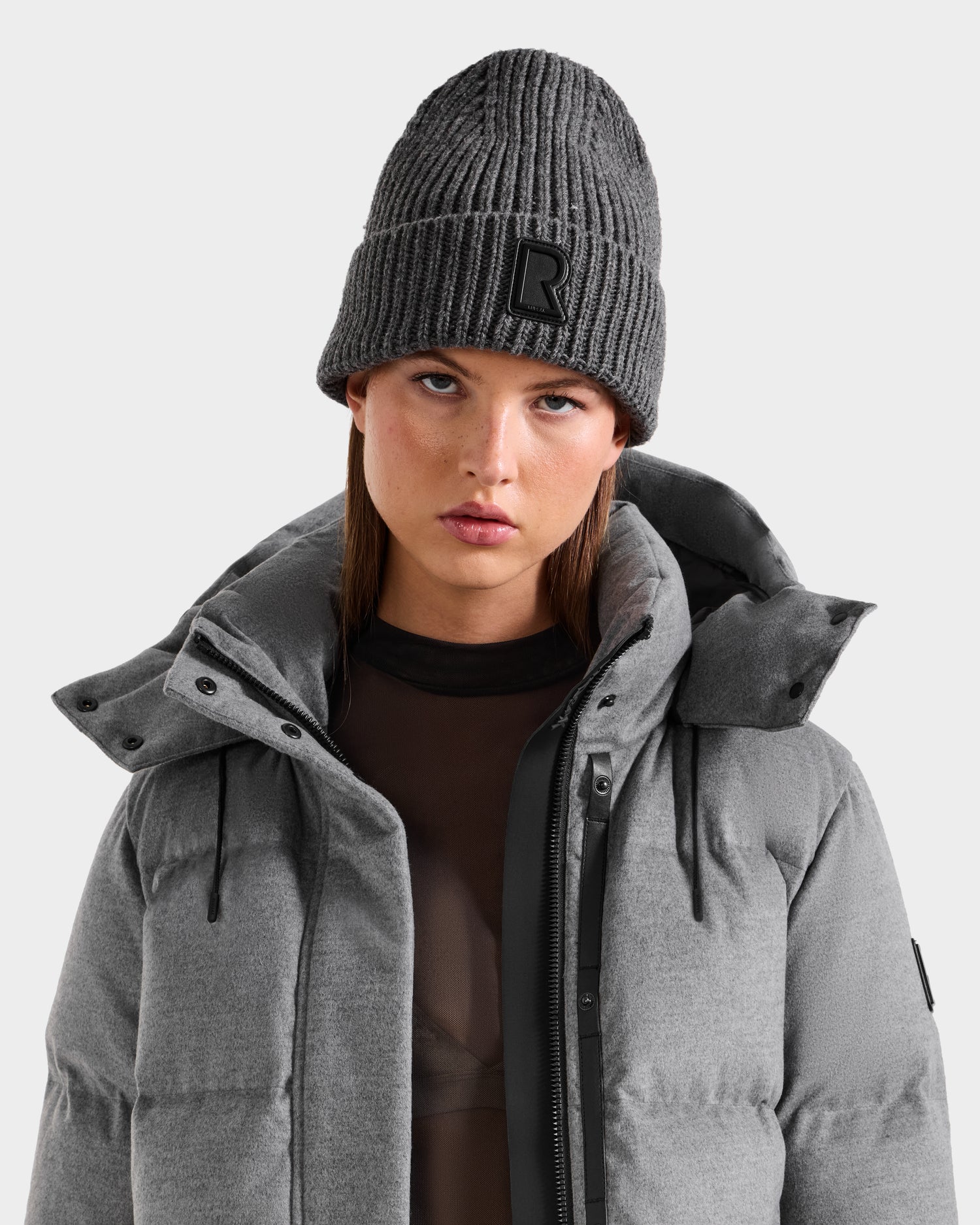 A woman in the CYBELLE - 9424504 Grey puffer jacket by Rudsak and a wool blend beanie with an R patch gazes forward with a neutral expression against a plain light background.