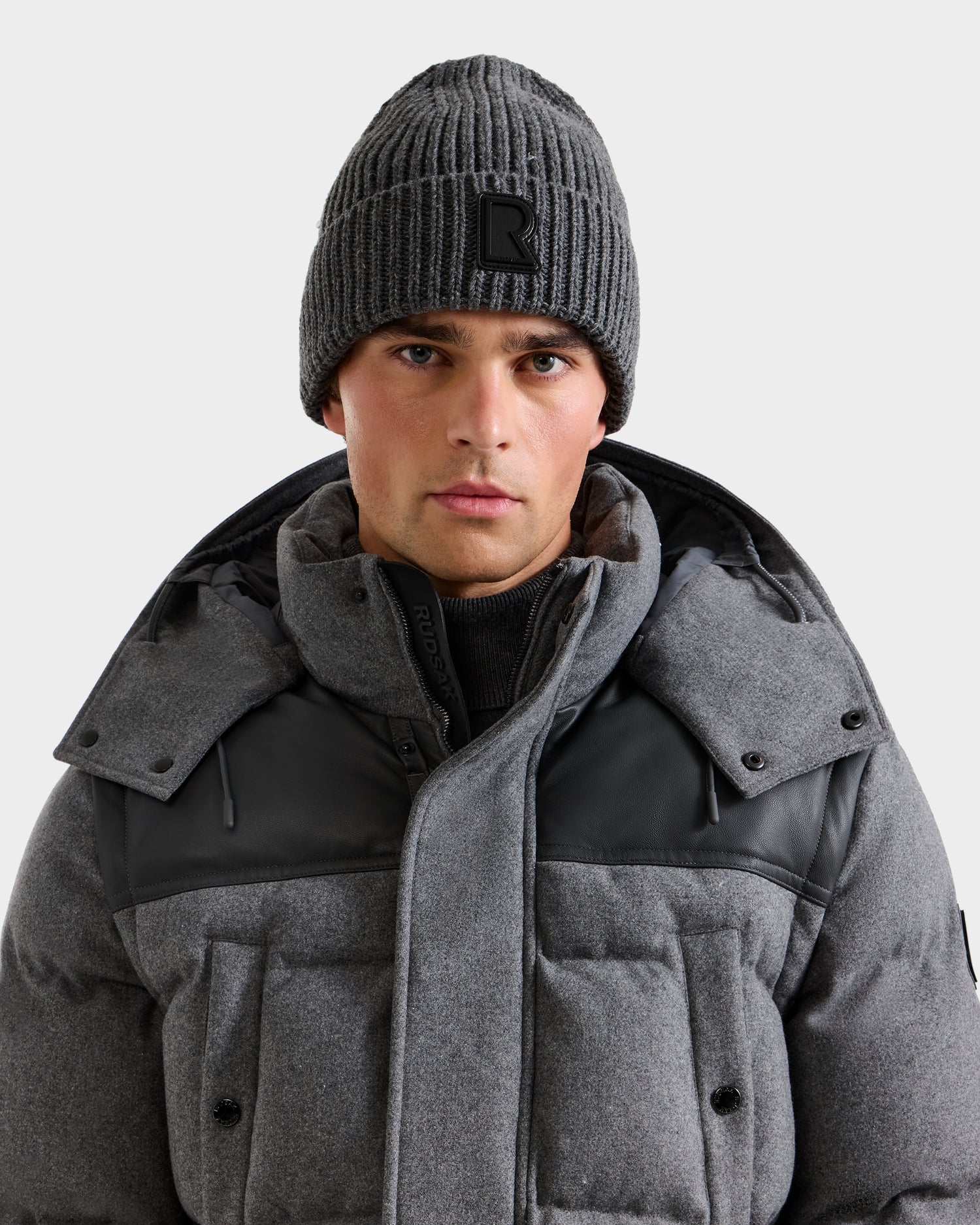 A man wearing the Rudsak CYBELLE - 9424504 Grey winter jacket and a gray wool blend beanie with a black patch, looking directly at the camera against a plain light background.