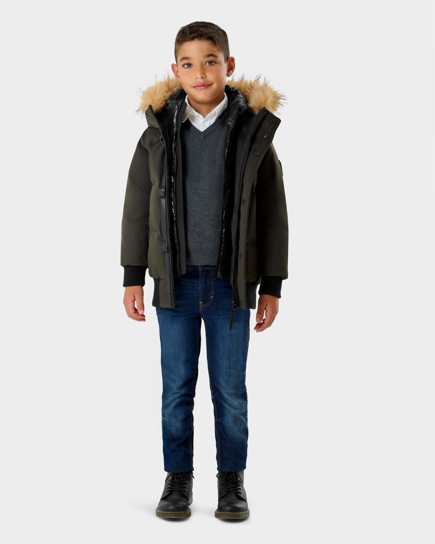 A young boy with short brown hair smiles slightly against a plain background, wearing the Rudsak DALLAS K - K125501 Dark Olive parka with a fur-lined hood, over a white shirt, gray sweater, blue jeans, and black boots.