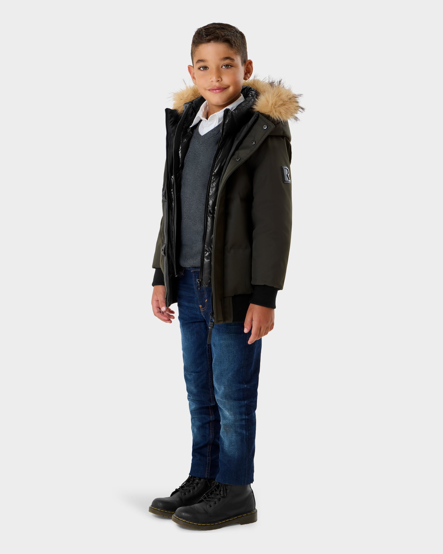 A young boy smiles in the Rudsak DALLAS K - K125501 Dark Olive parka with a fur-lined hood, gray sweater over a white shirt, blue jeans, and black boots, set against a plain light background.