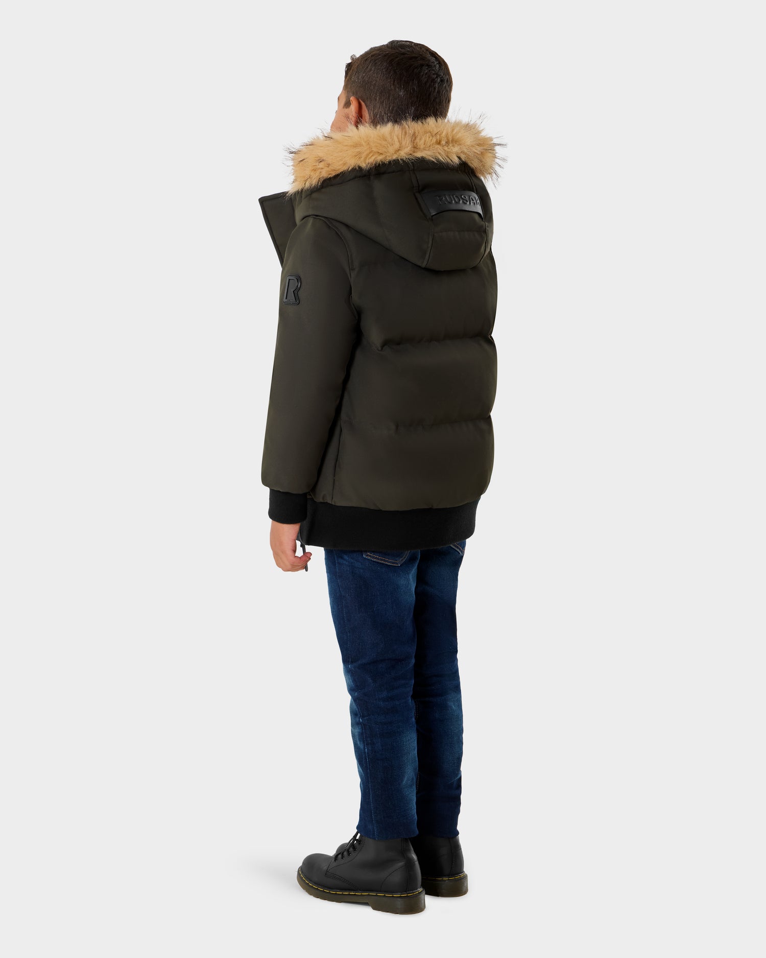 A boy faces away, wearing the Rudsak DALLAS K - K125501 Dark Olive puffer jacket with a fur-lined hood, paired with dark blue jeans and black boots against a plain light background.