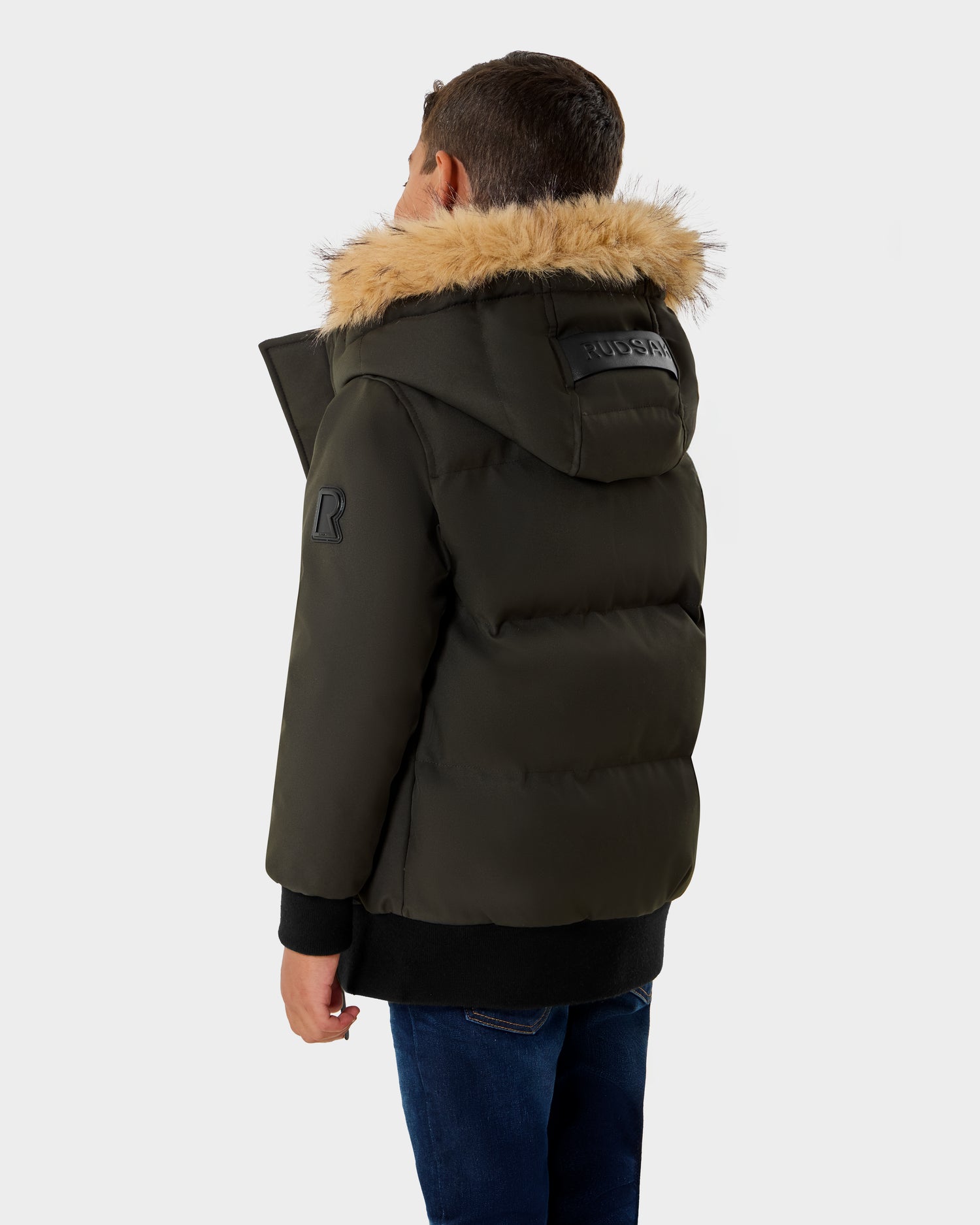 A young boy is seen from behind wearing the Rudsak DALLAS K - K125501 Dark Olive puffer jacket with a fur-lined hood and blue jeans, standing against a plain white background.