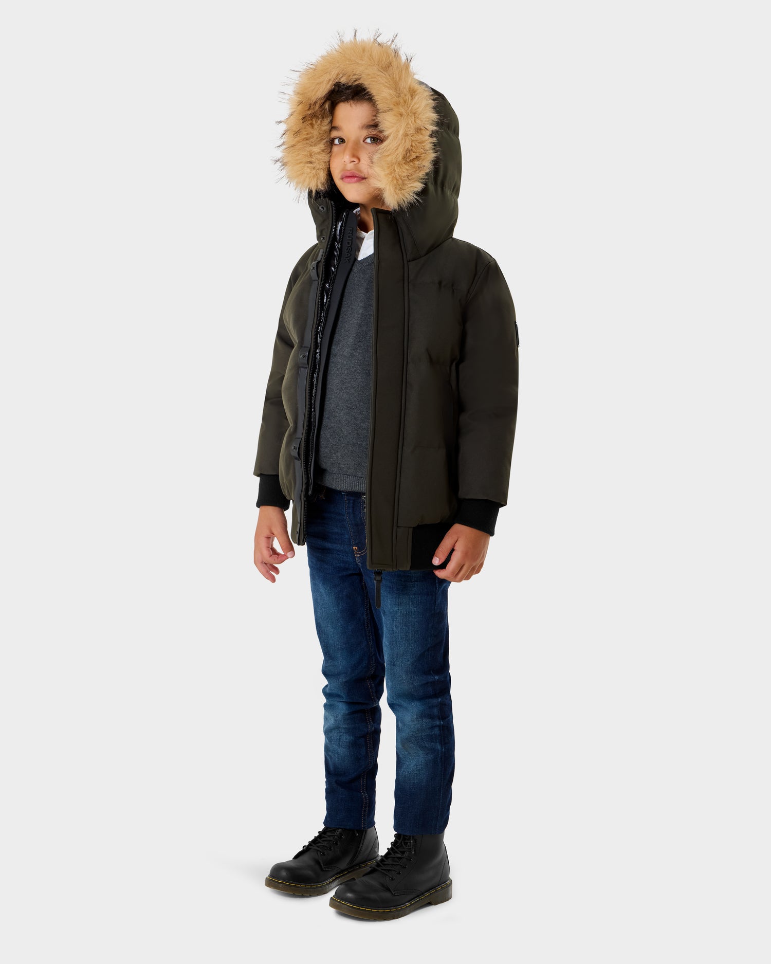 A young boy stands slightly to the side, wearing the Rudsak DALLAS K - K125501 Dark Olive hooded winter coat with fur trim, blue jeans, and black boots against a plain light background.