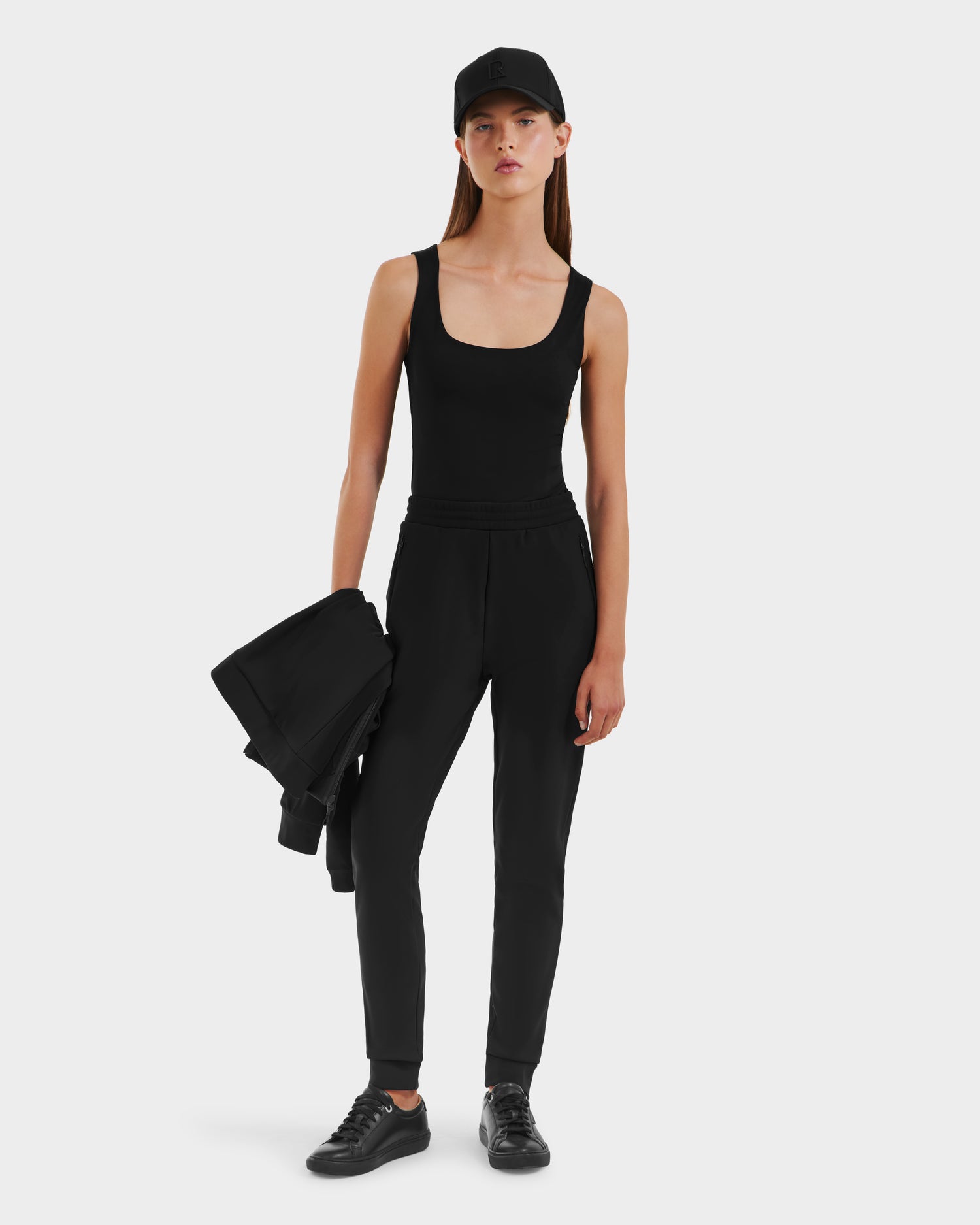 A woman in athleisure style stands against a white background, wearing the Rudsak DAMERIS - 8525575 Black tank top, jogger pants, black sneakers, a black cap, and holding a folded black jacket in her left hand.