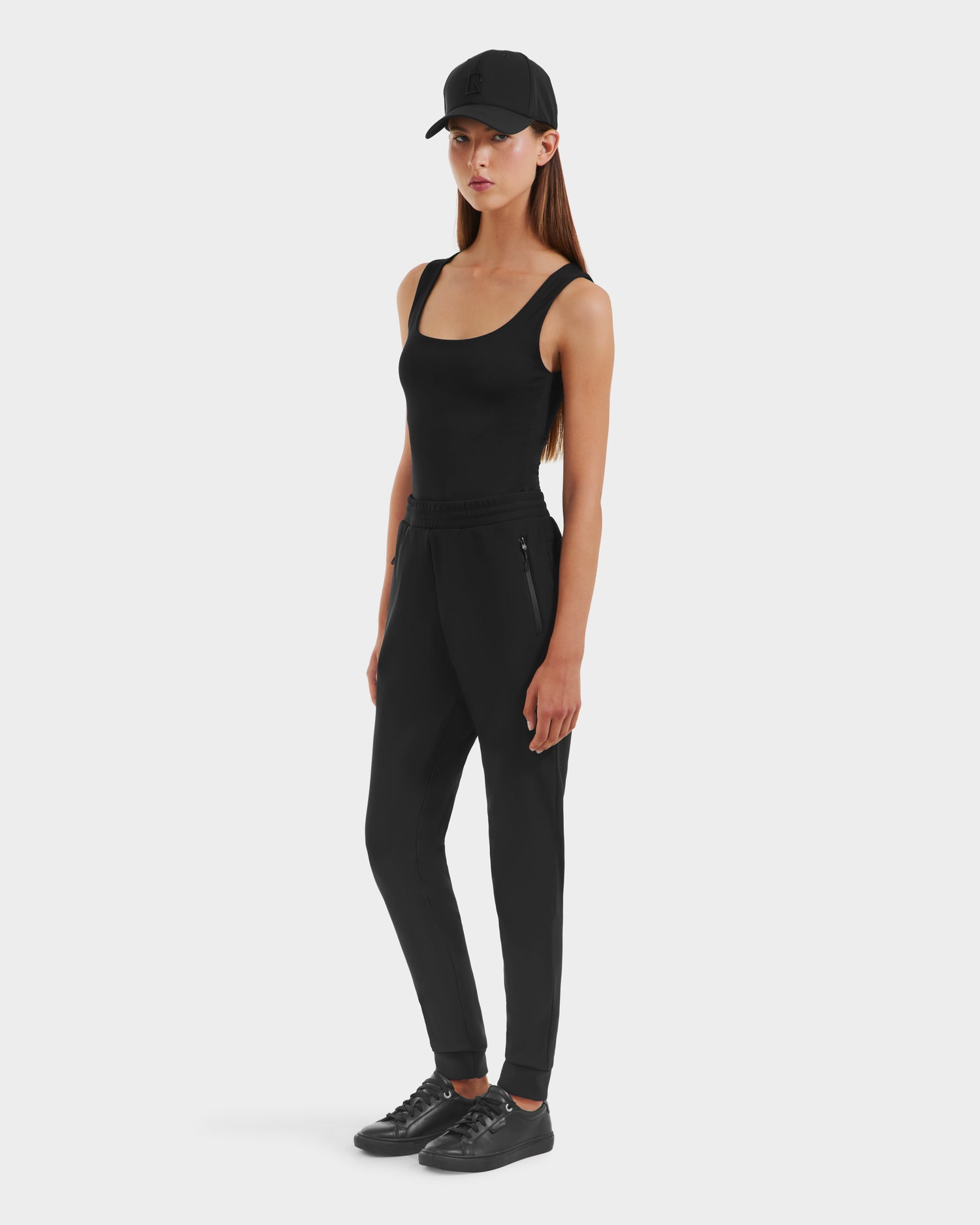 A woman poses against a plain background wearing Rudsak DAMERIS - 8525575 Black women’s jogger pants with a matching tank top, sneakers, and a baseball cap. Her long straight brown hair and neutral expression complete the chic athleisure look.